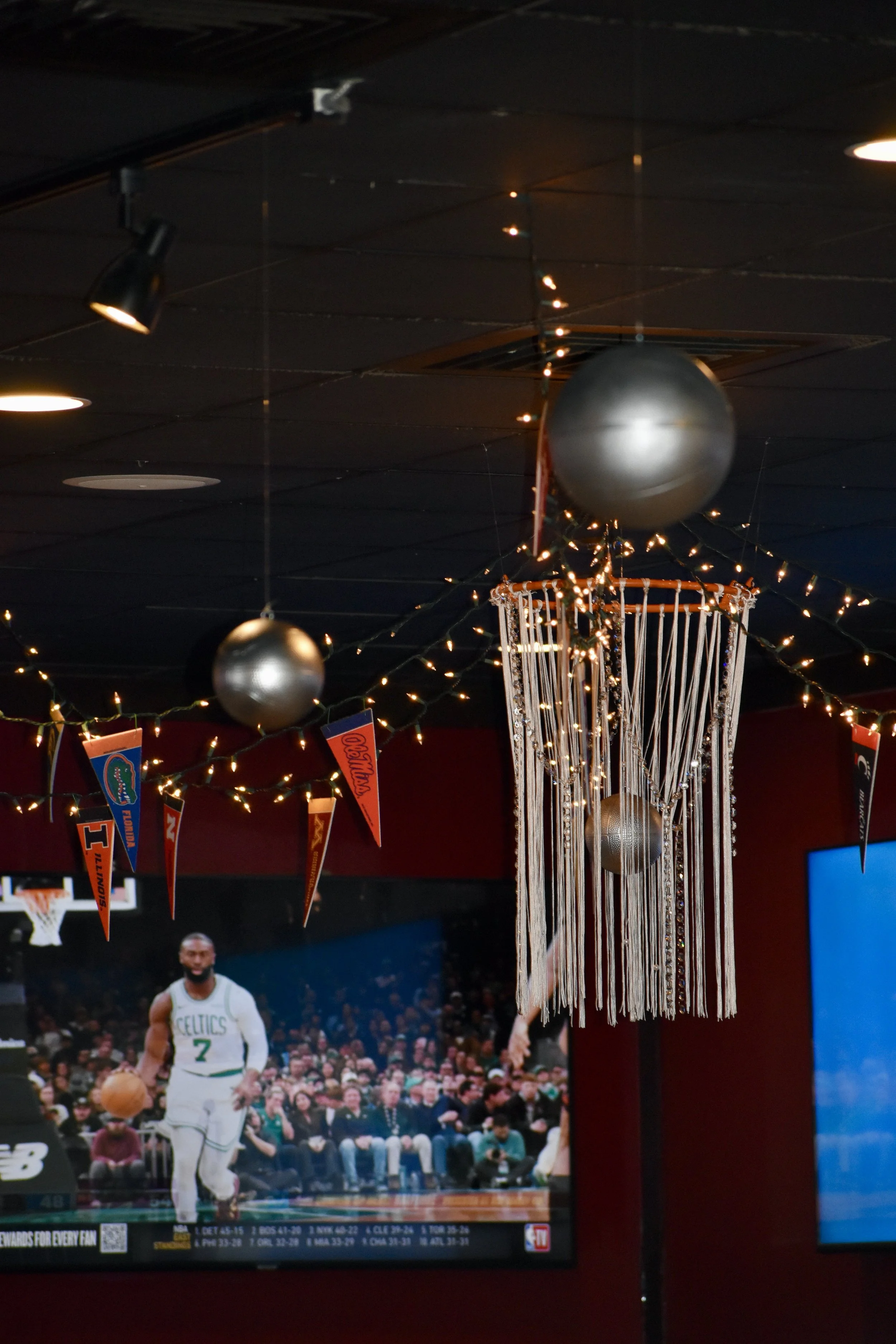 Decorative basketball-themed decor with hanging metallic spheres, string lights, and a banner displaying the University of Florida and Illinois flags, in an indoor sports bar. March Madness