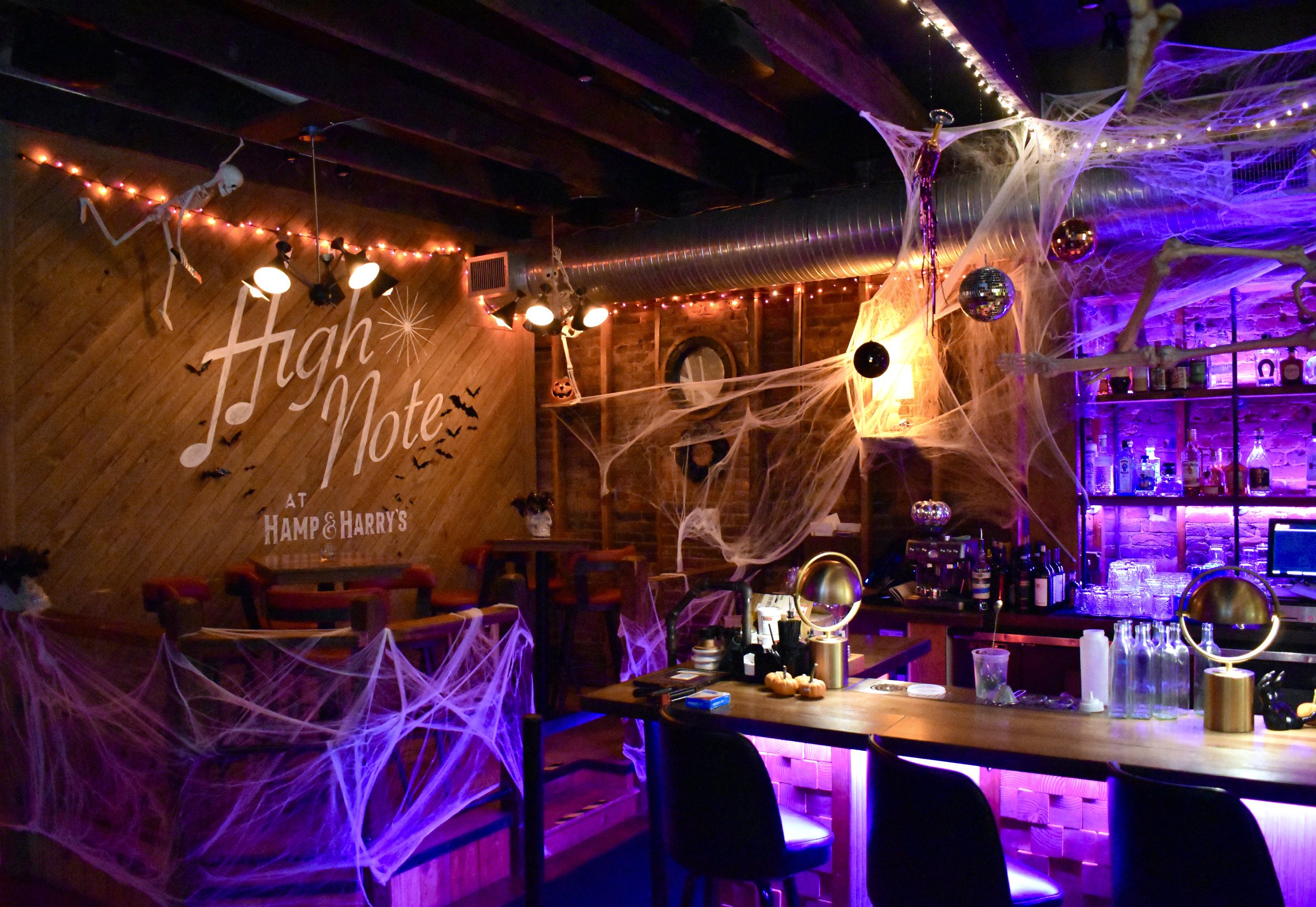 Decorated bar area with Halloween decorations including spider webs, skeletons, and pumpkins, with purple and orange lighting. Disco themed halloween party.