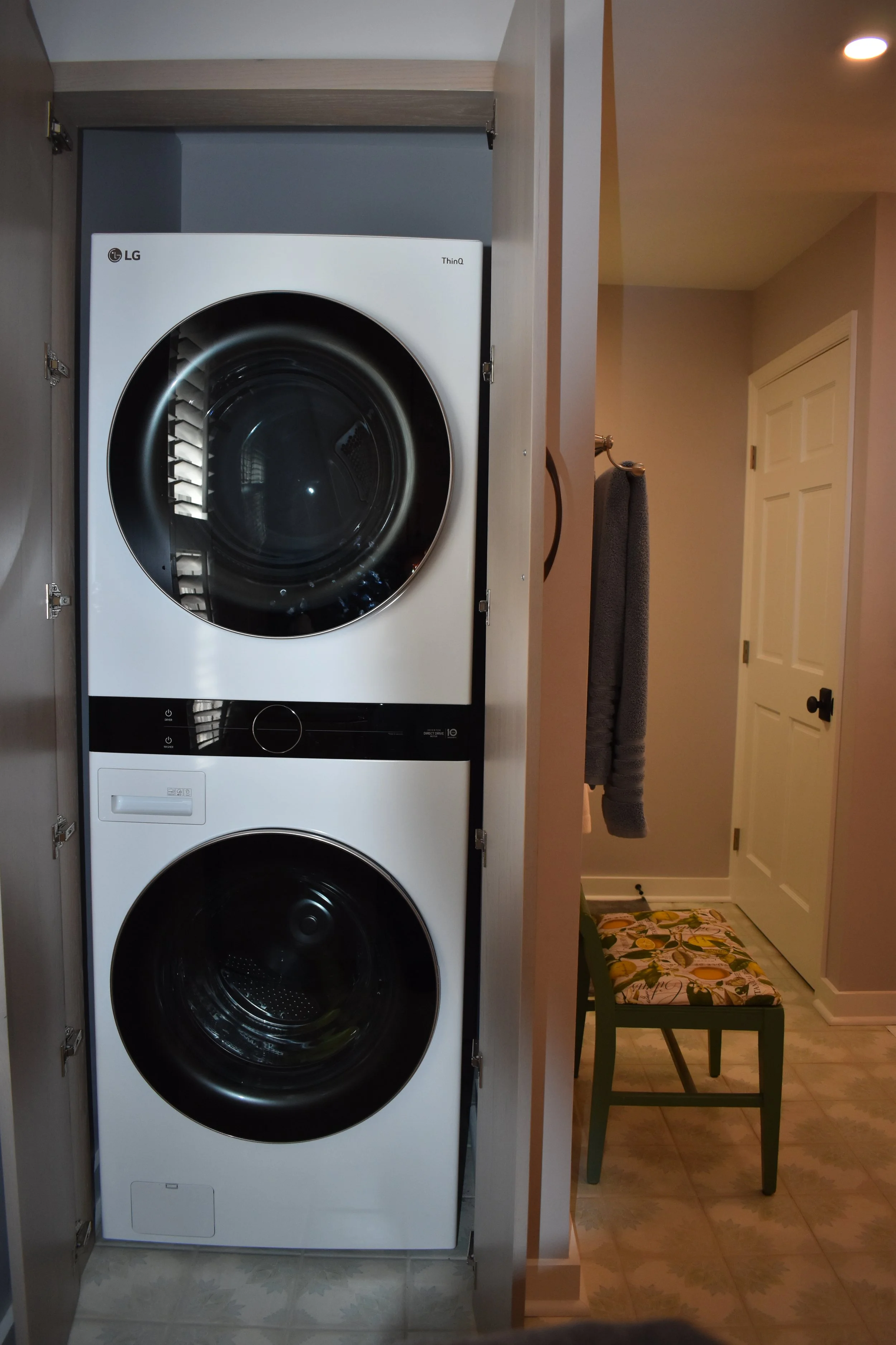 A stacked LG washer and dryer inside a closet. The closet is open, showing the appliances, with a gray towel hanging on a hook nearby and a small green bench with a floral cushion in front of a white door.