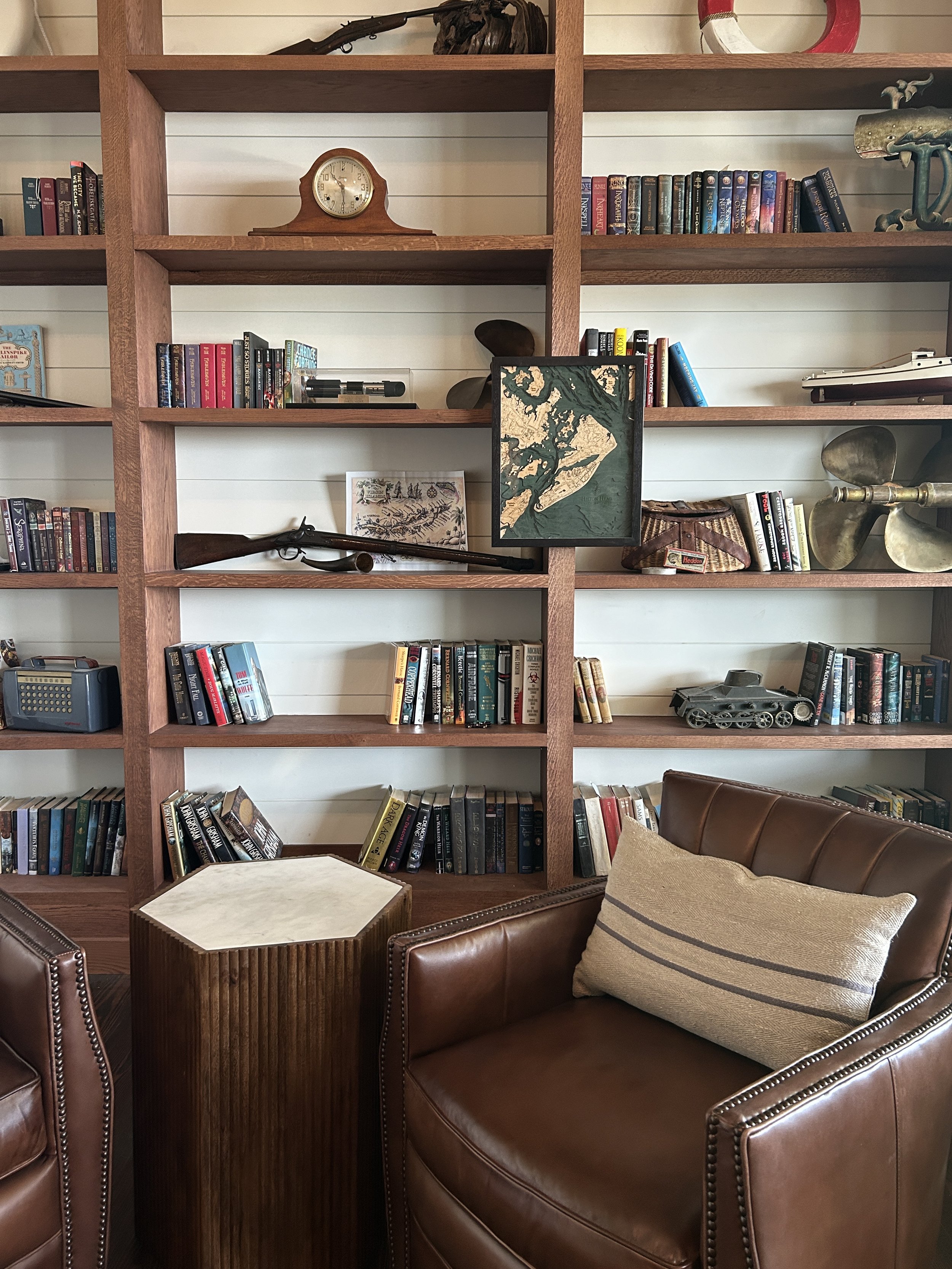 A wooden bookshelf filled with books and decorative items, including a clock, framed artwork, a rifle, and a model tank, with leather chairs and a small wooden side table in front. Hilton Head Island Interior Design.