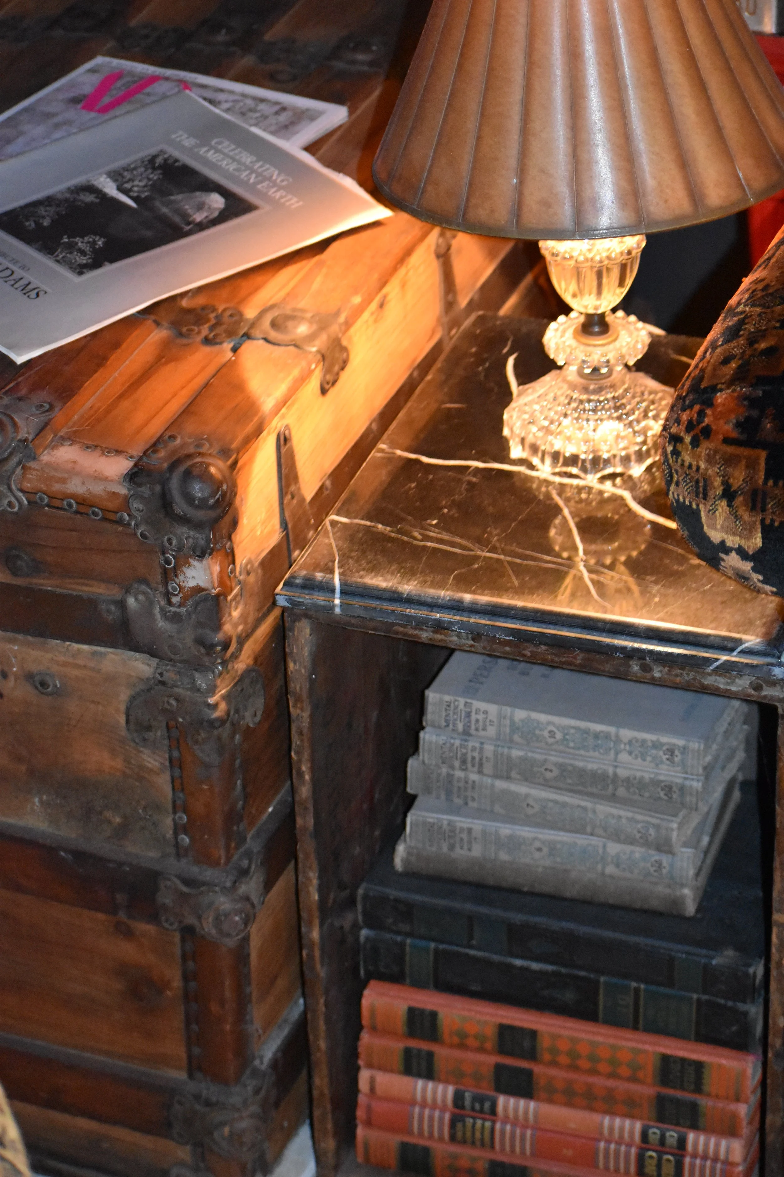A wooden chest and a black bookshelf holding vintage books under a table lamp with a brown shade.