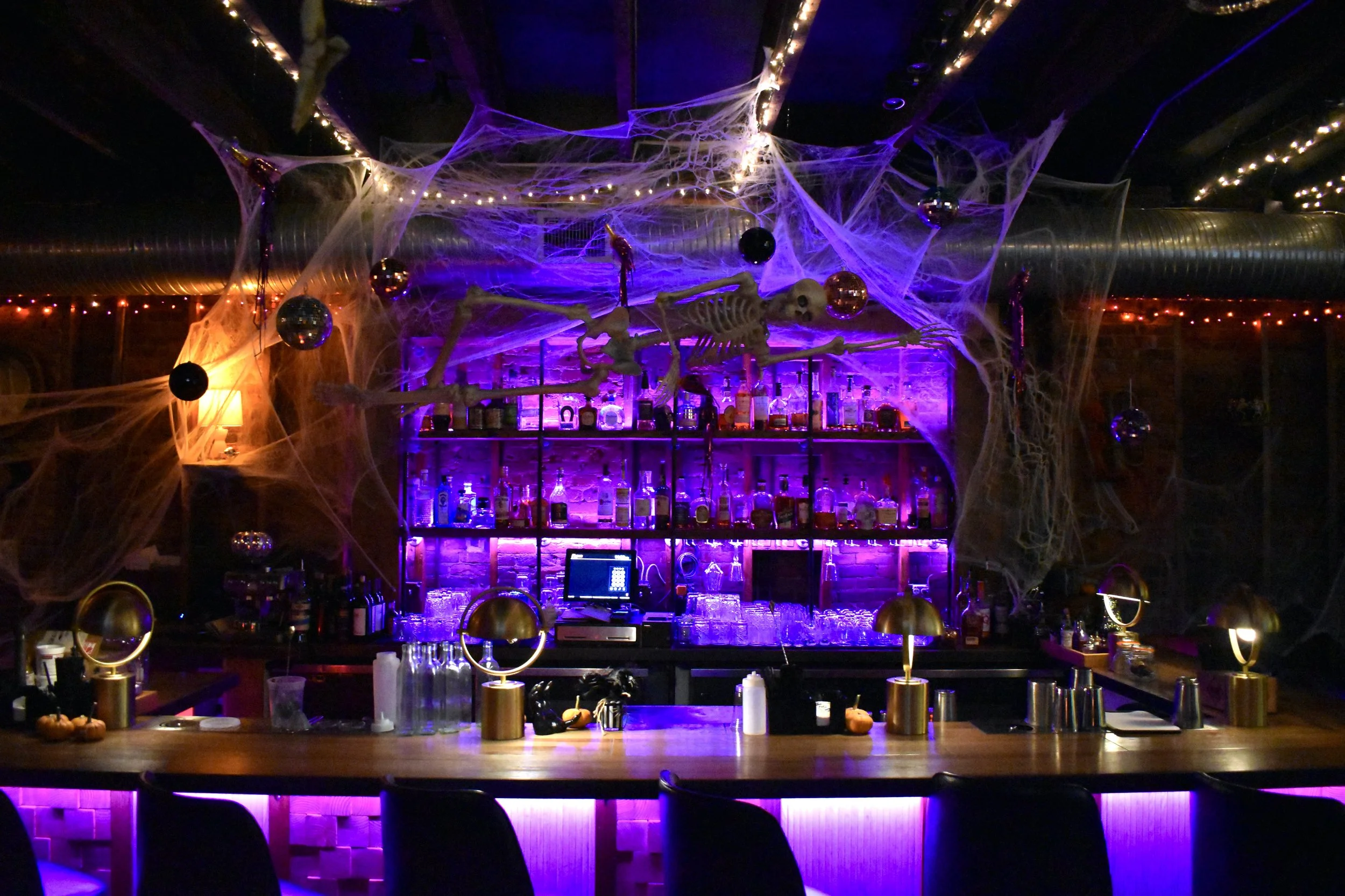 A bar decorated for Halloween with spider webs, Halloween ornaments, and a skeleton skeleton hanging above the bar. The bar is lit with purple and orange lights, with a variety of bottles and glasses on the shelves. Halloween-themed decorations are o