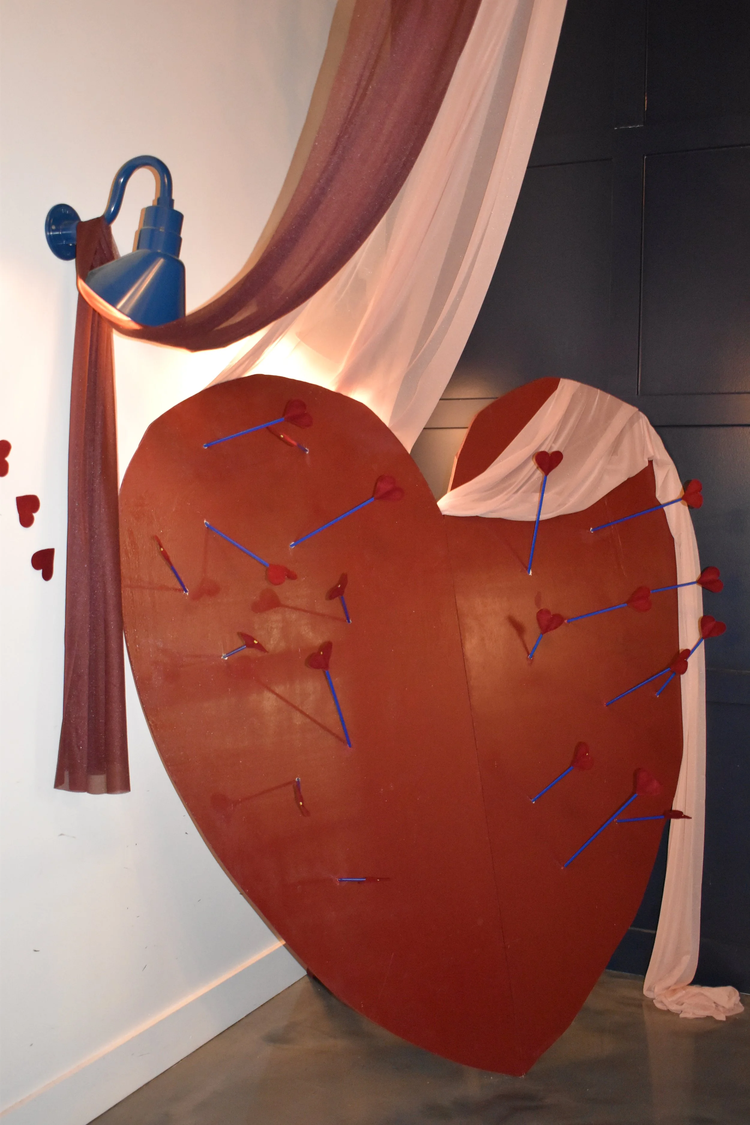 A large wooden heart-shaped board decorated with multiple small red heart-shaped decorations on blue sticks, placed against a wall with curtains and a wall-mounted lamp.