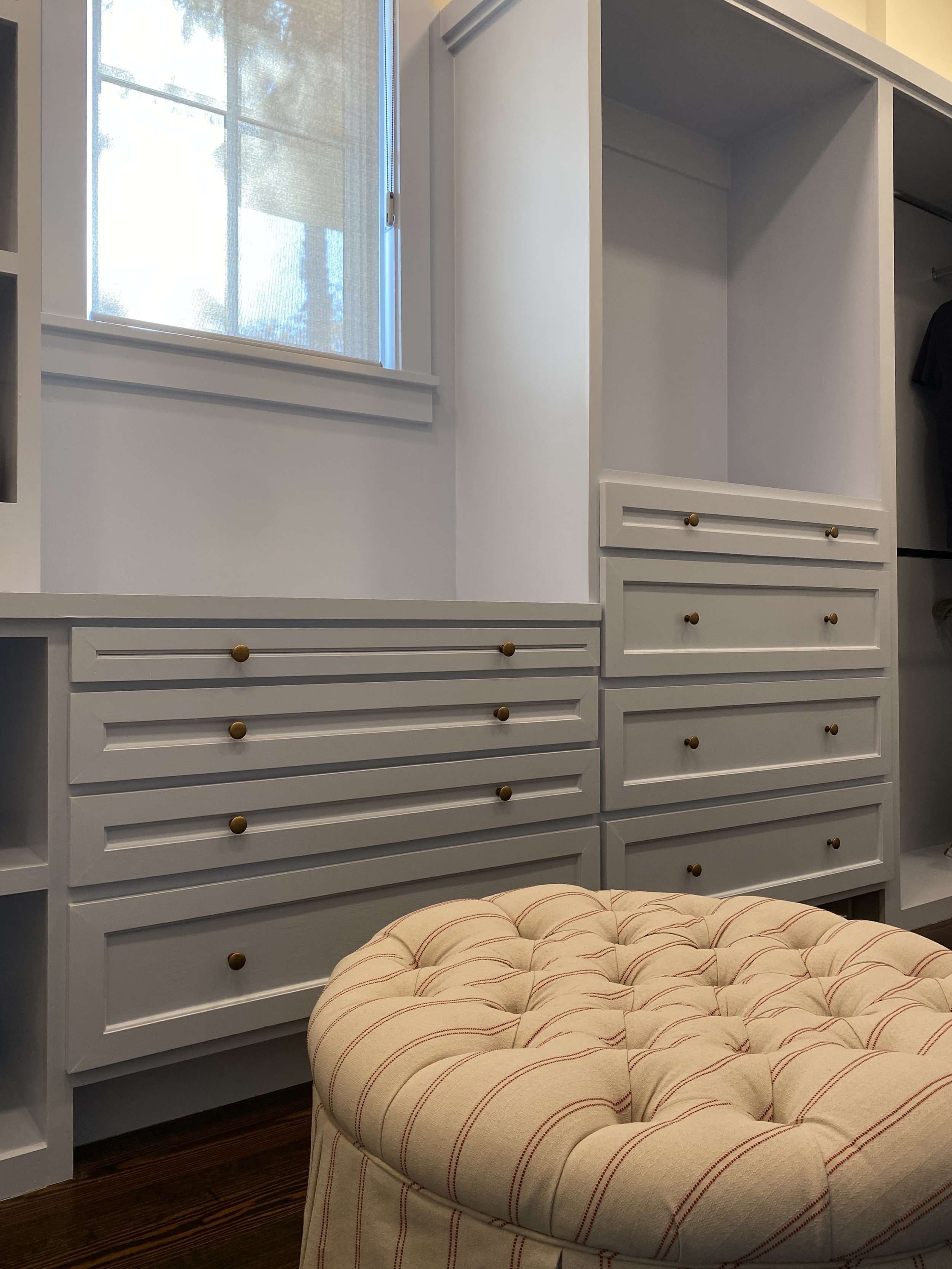 A custom closet with white built-in drawers and an open shelving unit, a window with a sheer curtain, and a beige upholstered ottoman with tufted buttons in the foreground. Hilton Head Island Interior Design.