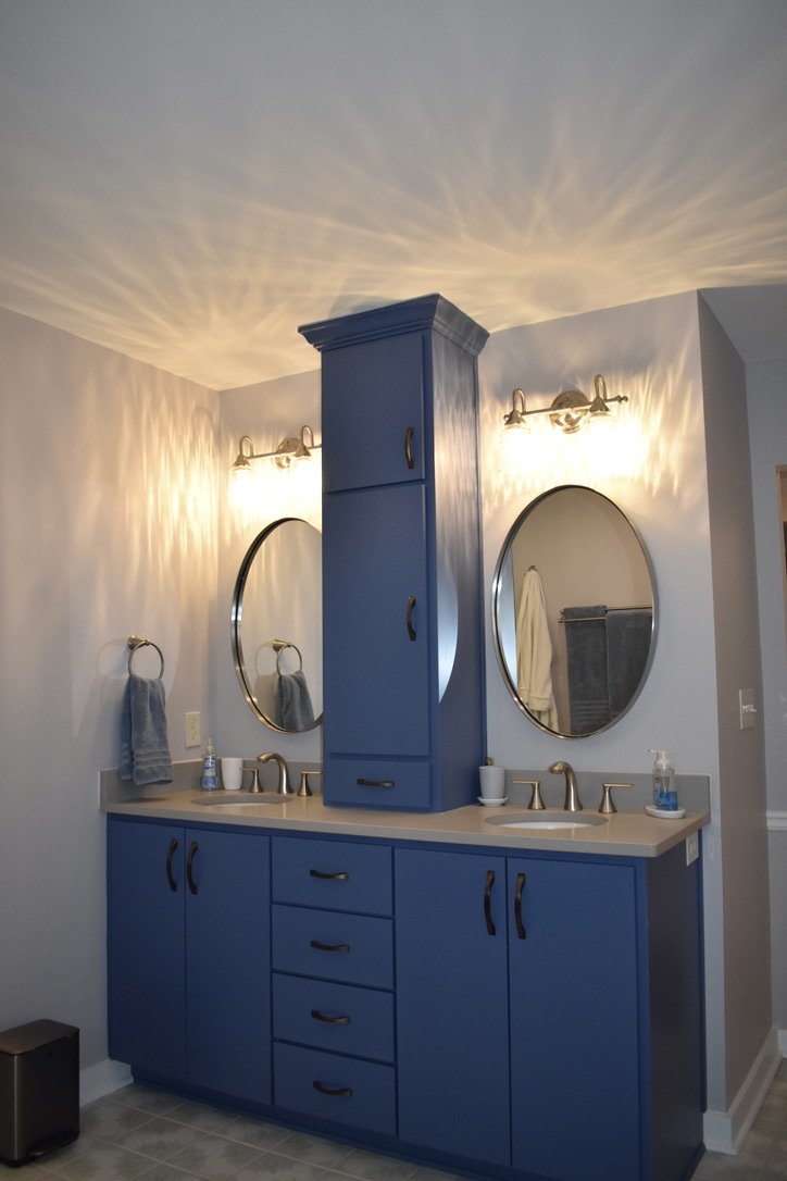 Bathroom with blue cabinets, two oval mirrors, and two wall-mounted light fixtures. The sinks have gold-colored fixtures, and there are towels and bathroom accessories on the counter.