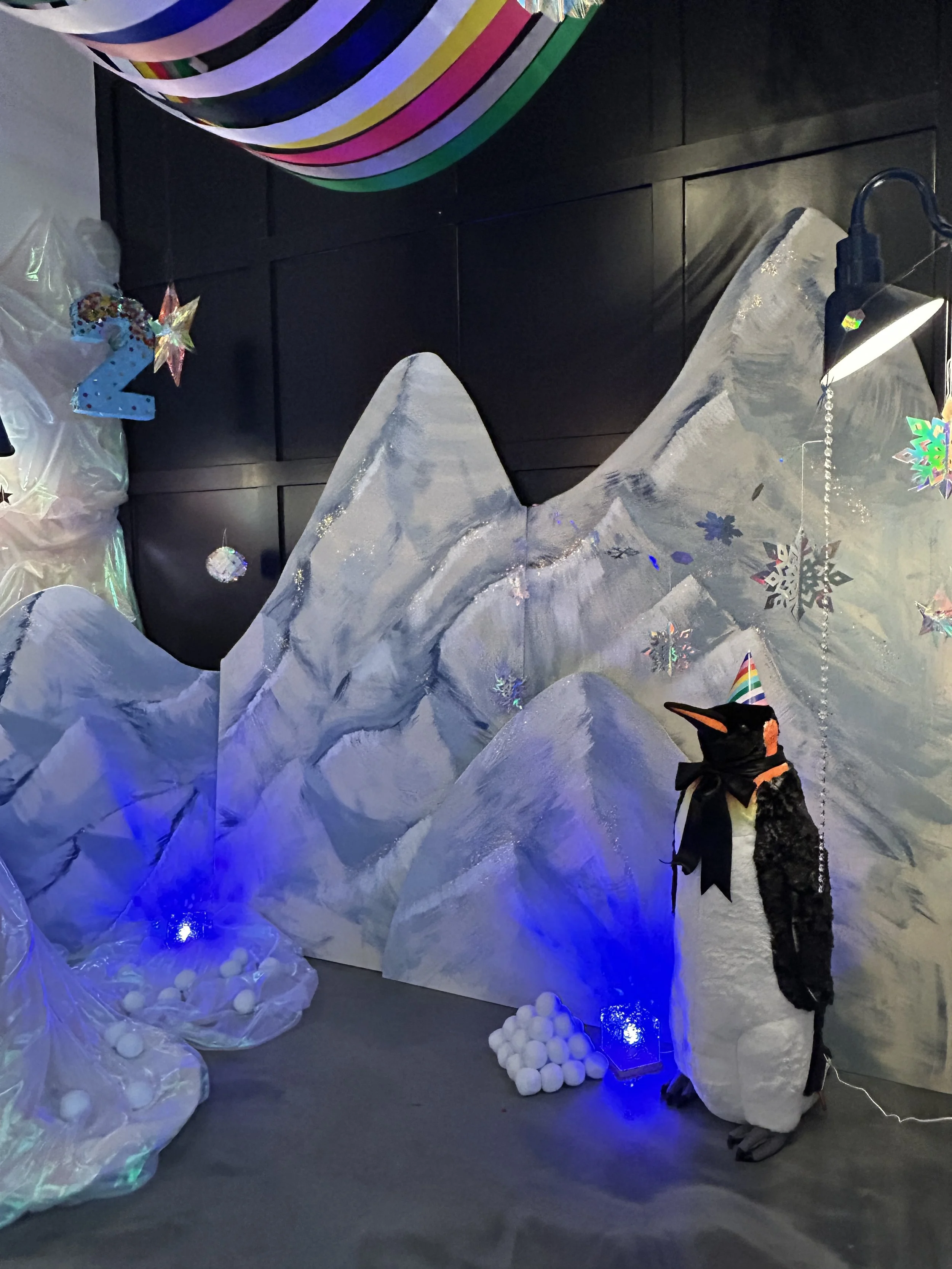 Decorated scene with artificial snow mountains, a penguin plush wearing a rainbow party hat and black scarf, snowflake decorations, colorful hanging stripes, and a blue number 2 balloon, likely part of a winter or holiday themed setup.