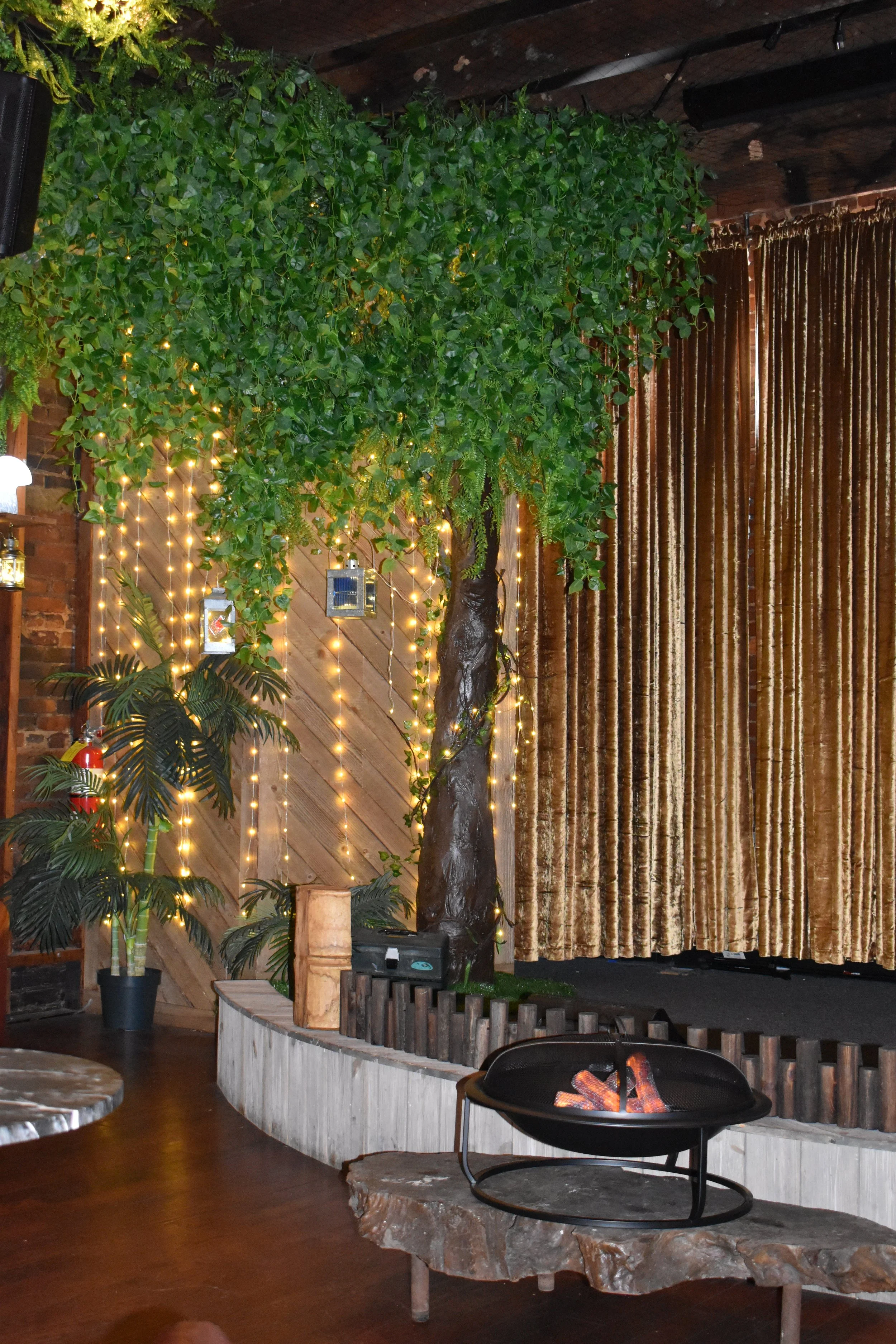 Indoor scene featuring artificial trees decorated with string lights, gold curtains, potted plants, and a small fire pit on a stone table. Hamp & Harry's Marietta Square.