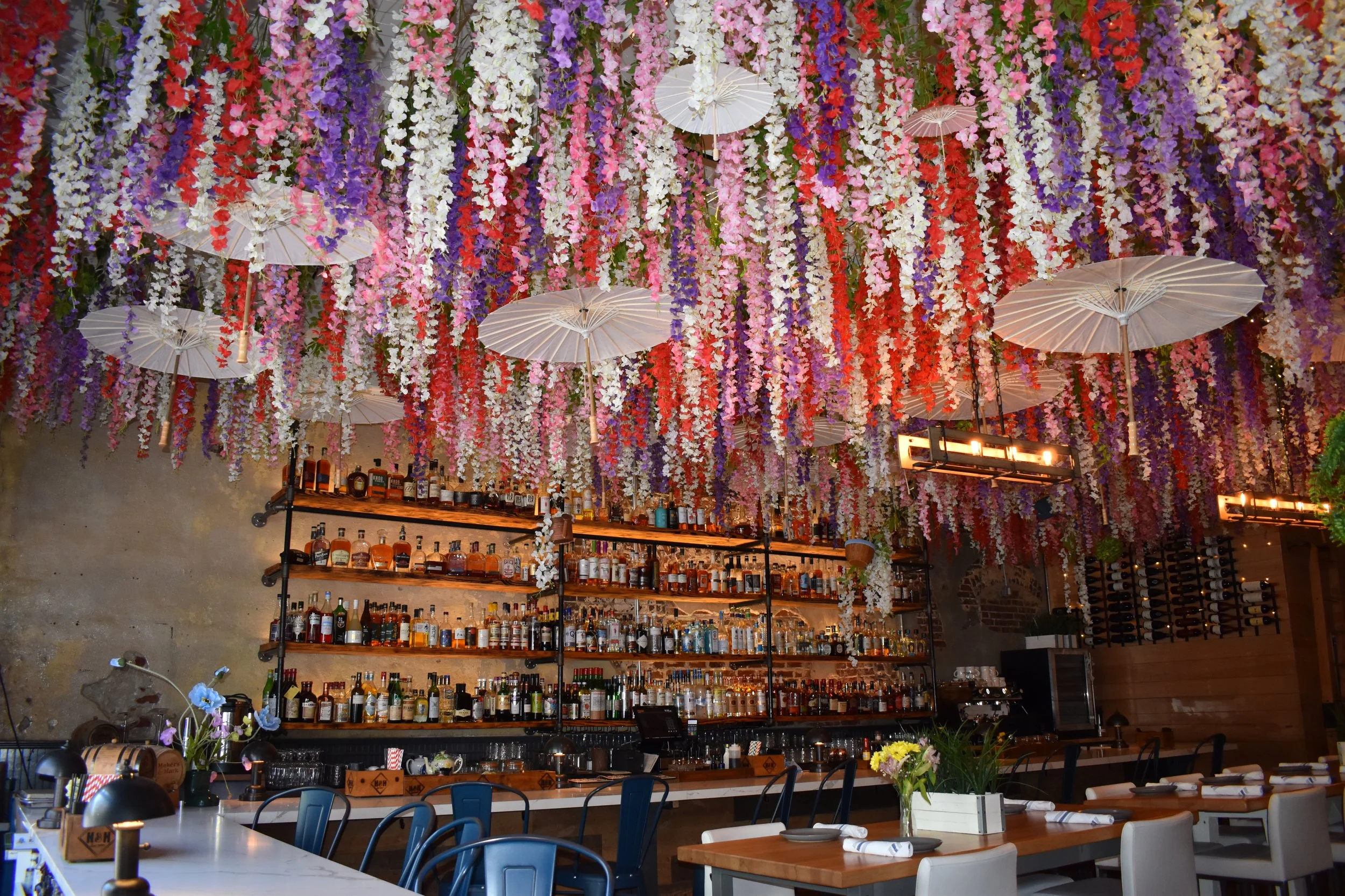 Interior of a restaurant or bar decorated with a ceiling of hanging colorful flowers and white paper umbrellas. Shelves behind the bar stocked with various bottles of alcohol and glasses. Tables set with white napkins and flowers.