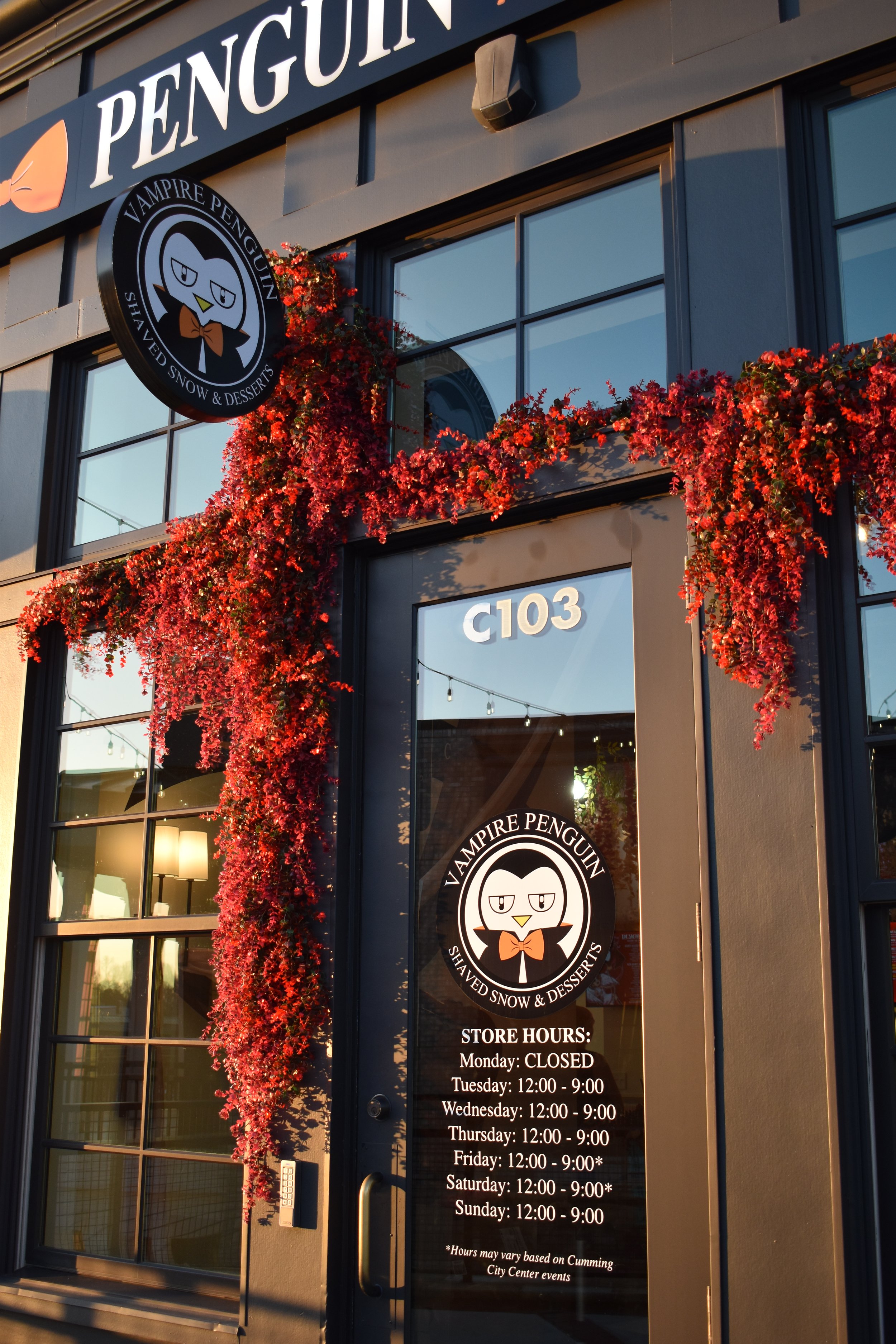 Storefront of Vampire Penguin, an ice cream and dessert shop, with red flowering plants hanging over the entrance, a sign with a penguin logo, and store hours listed on the door.