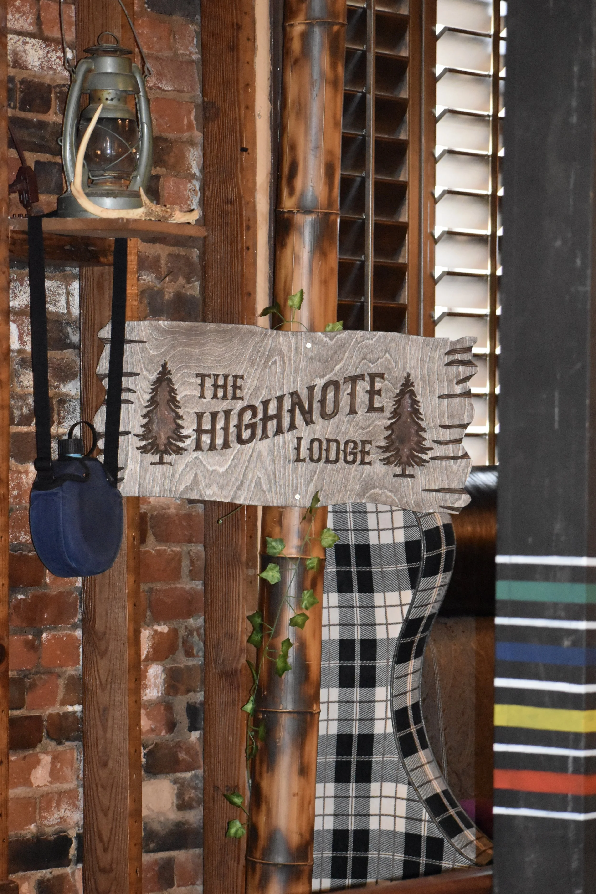 Wooden sign reading 'The Highnote Lodge' with trees engraved, hanging on a wooden post in a rustic interior with exposed brick wall and a mannequin wearing plaid clothing. Hamp & Harry's Marietta Square.