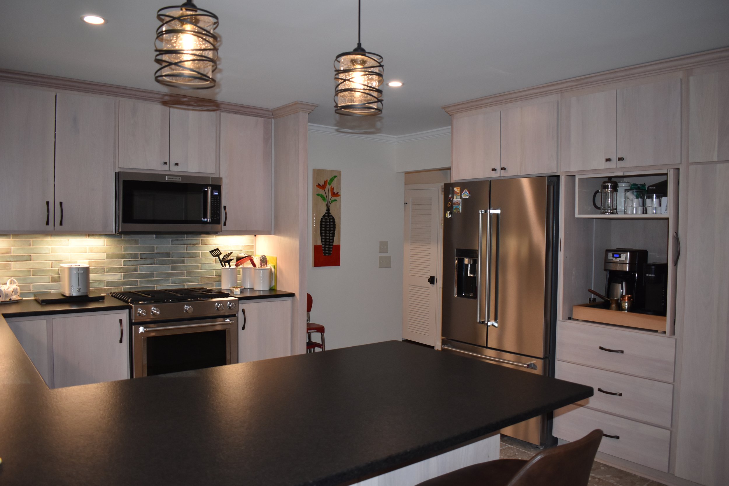 Modern kitchen with light wood cabinets, stainless steel refrigerator, black countertop, and tile backsplash. Two pendant lights hang from the ceiling, and a microwave is mounted above the stove. A coffee maker and glass jars are on the countertop. 