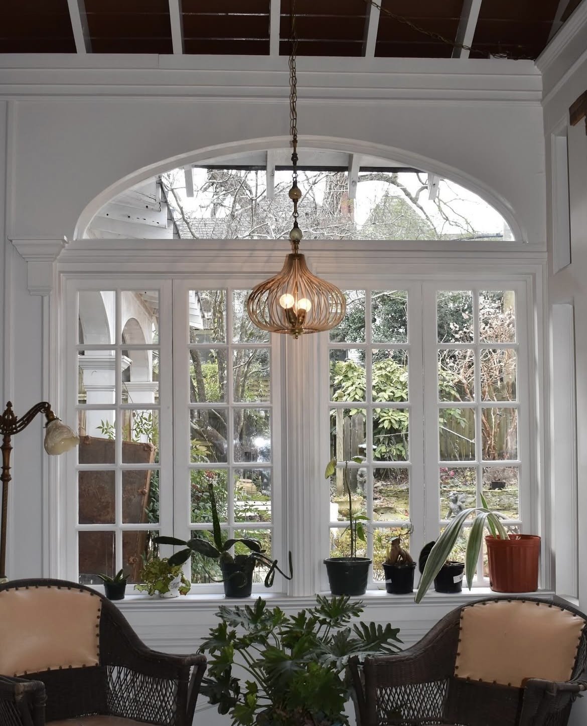 View of a sunroom with large white-framed windows and a view of a garden outside, featuring potted plants on the windowsill and a custom hanging light fixture. Custom swag lamp made from reclaimed lamp parts.