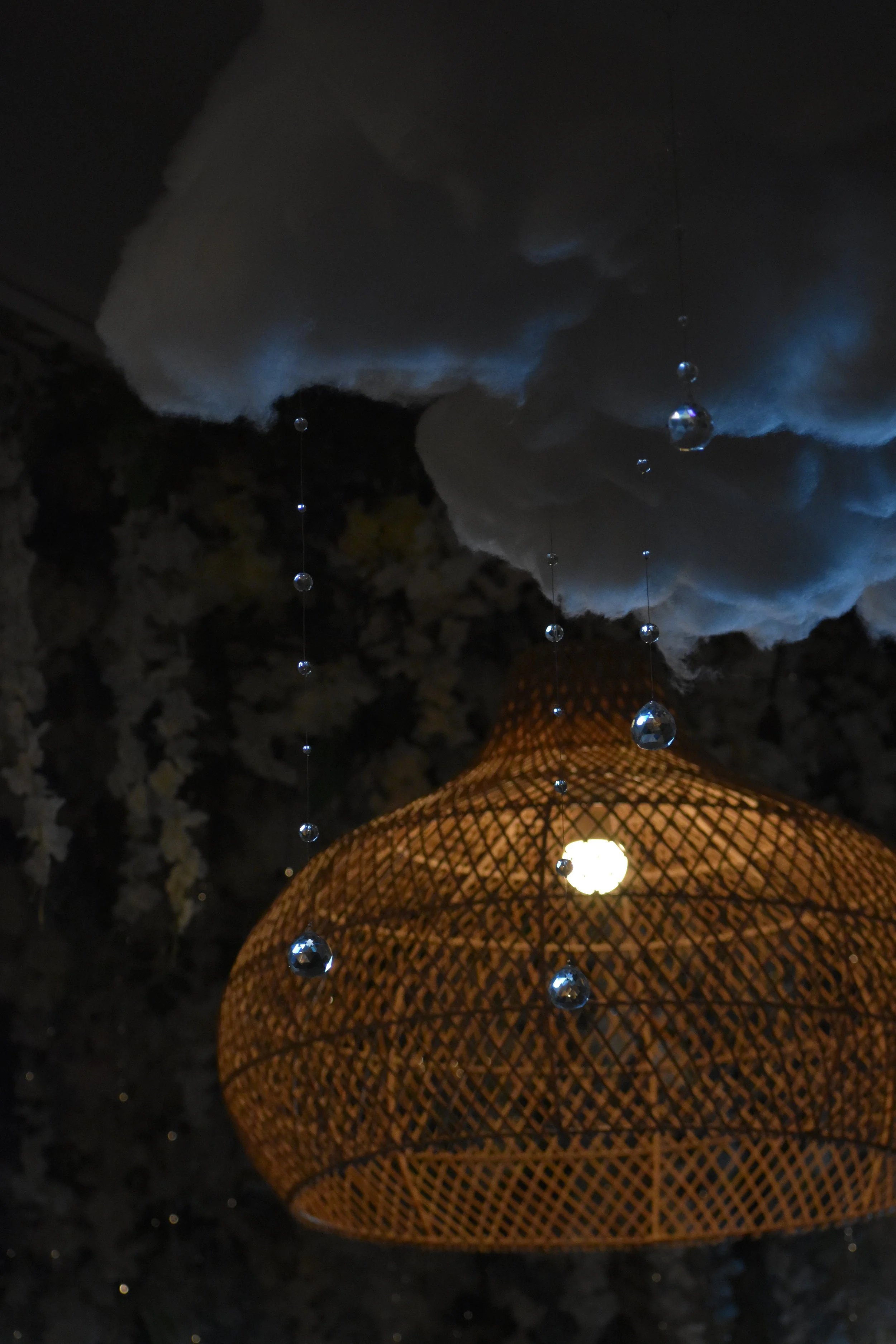 A woven rattan pendant lamp hanging from the ceiling, decorated with strands of small crystal beads, with a cloud-shaped fabric ceiling decoration above.