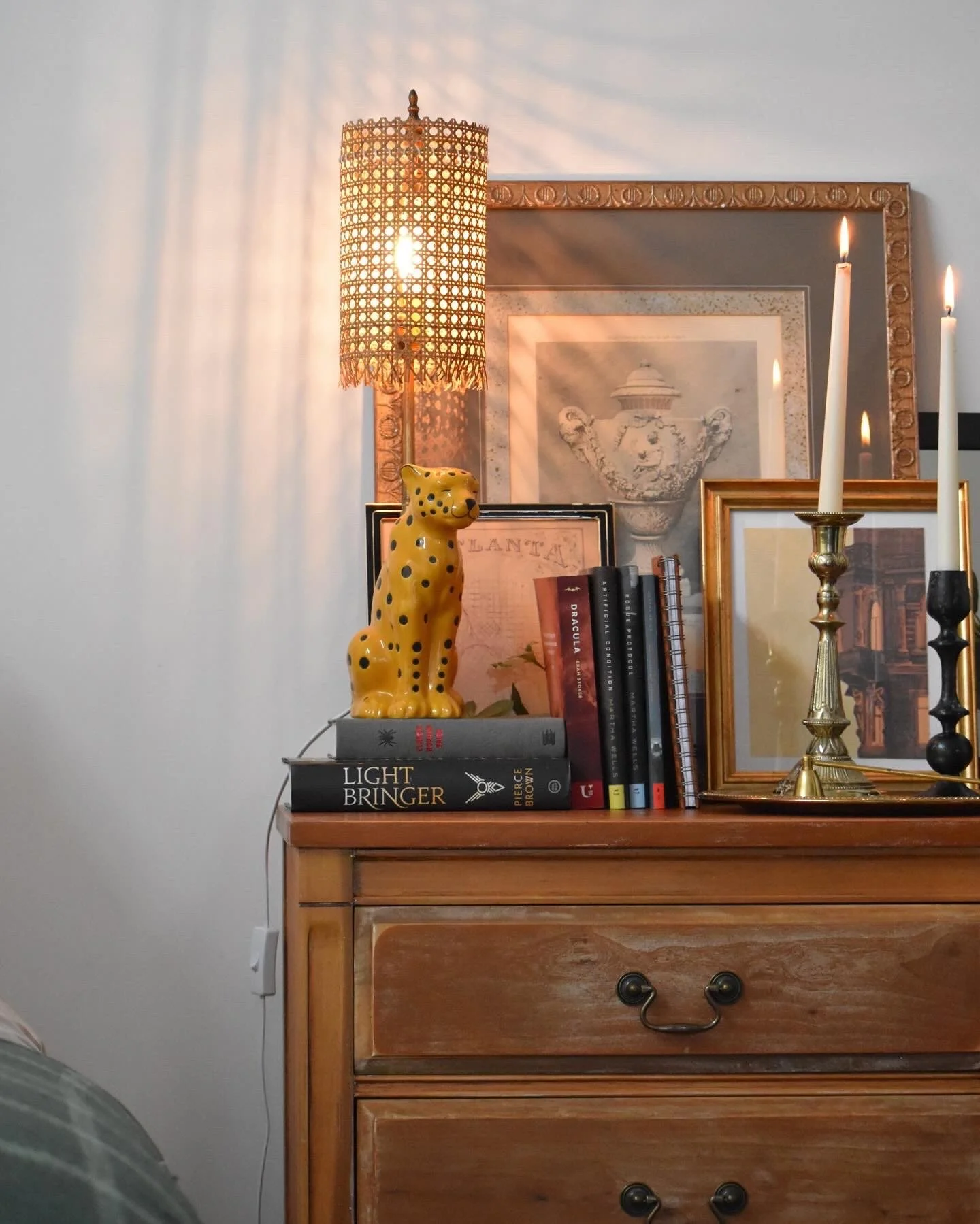 A wooden dresser with decorative items including a yellow leopard figurine, books, a framed picture, a mirror, and candles in a gold and black candlestick, with a woven lampshade casting light and shadows on the wall.