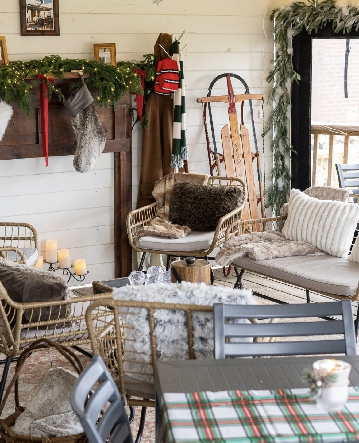 Cozy living room decorated for Christmas with a fireplace mantel adorned with greenery, lights, stockings, and framed pictures, alongside vintage sleds and warm seating with pillows and blankets.