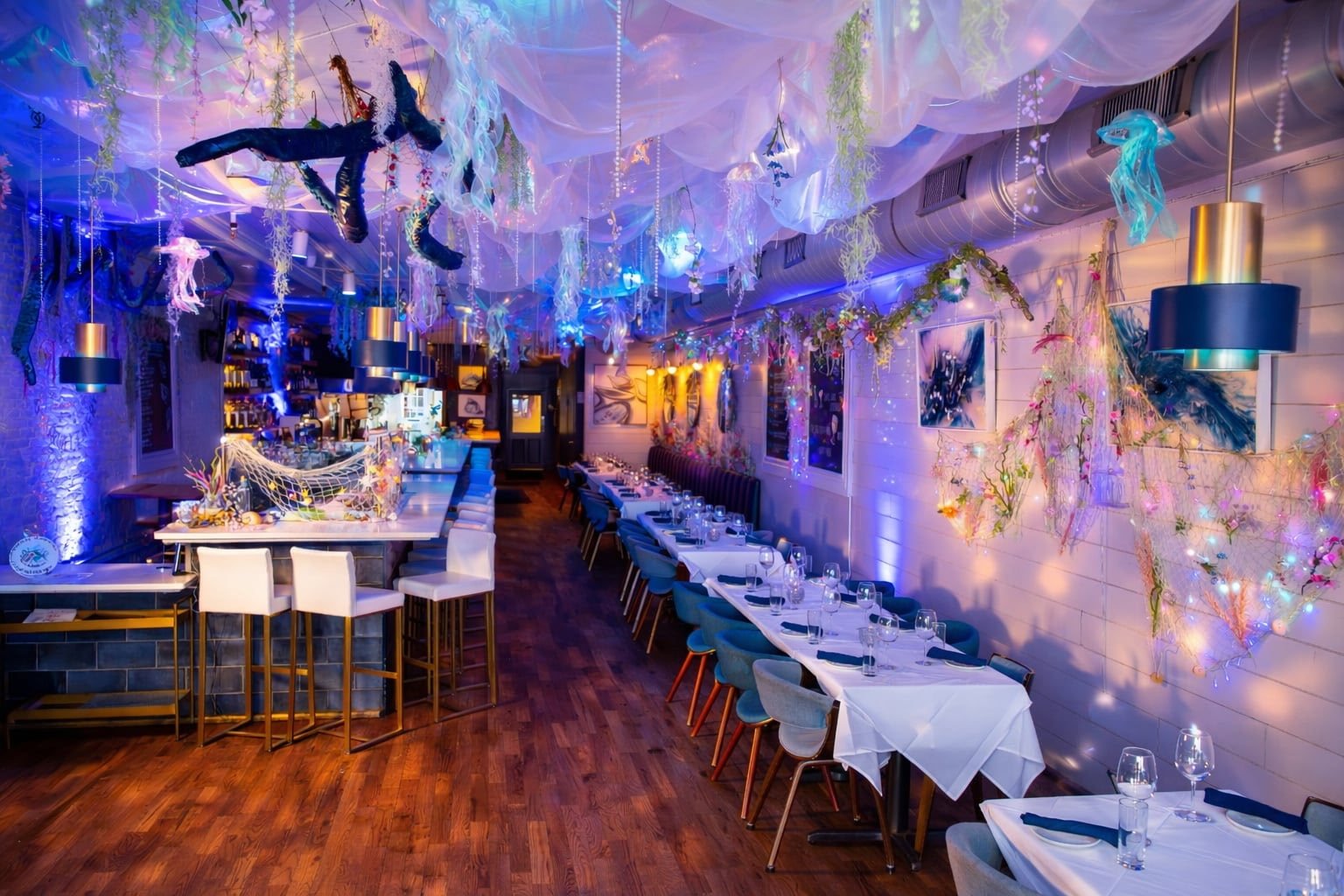 Interior of a restaurant decorated with colorful, ocean-inspired decor, including hanging jellyfish and fish, soft lighting, and tables set with white tablecloths and glasses.