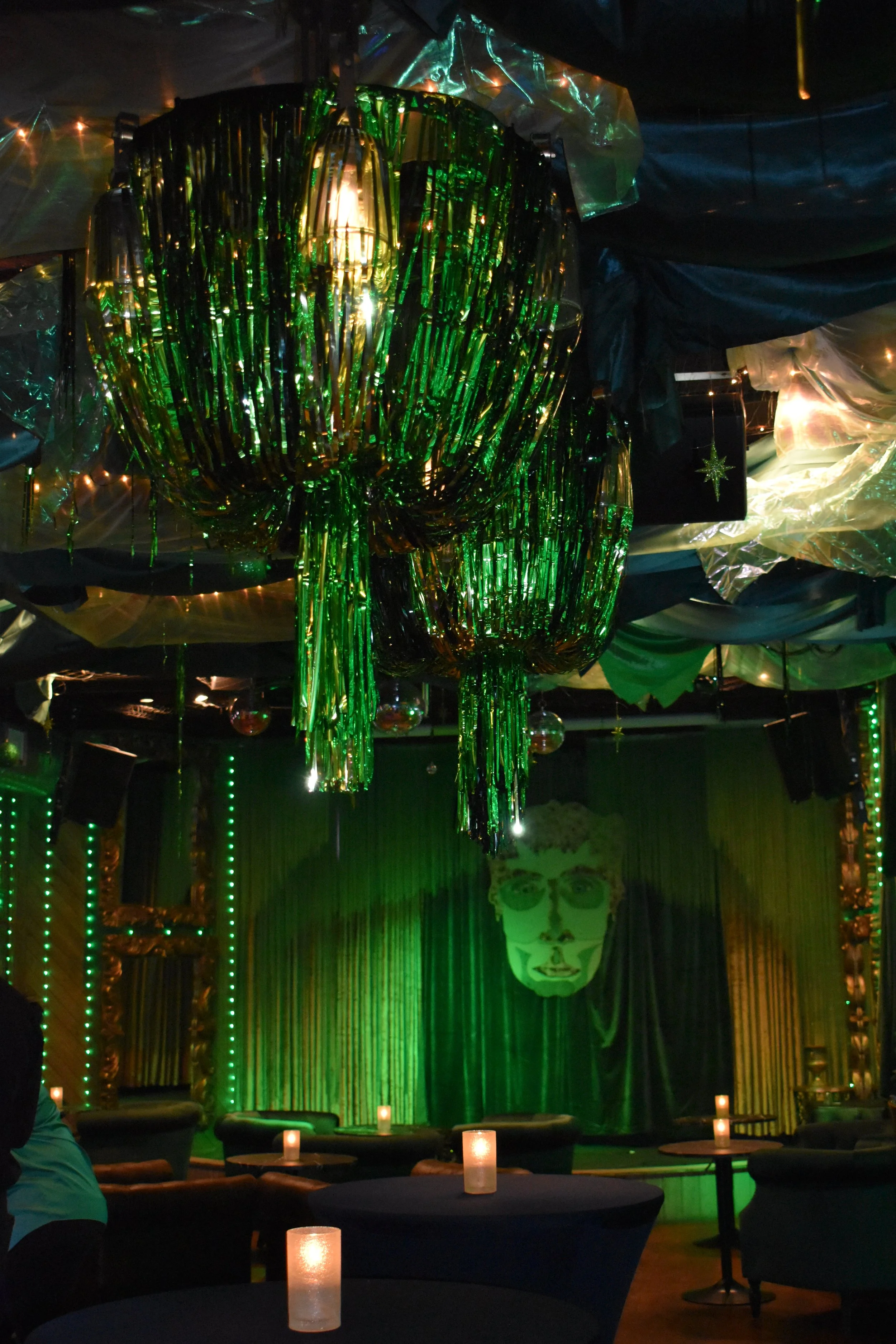 Indoor lounge decorated with green and gold curtains, hanging green and gold tinsel chandeliers, and small candles on tables, with a large stylized face portrait on a curtain in the background. Wizard of Oz themed decor featuring Emerald City. 
