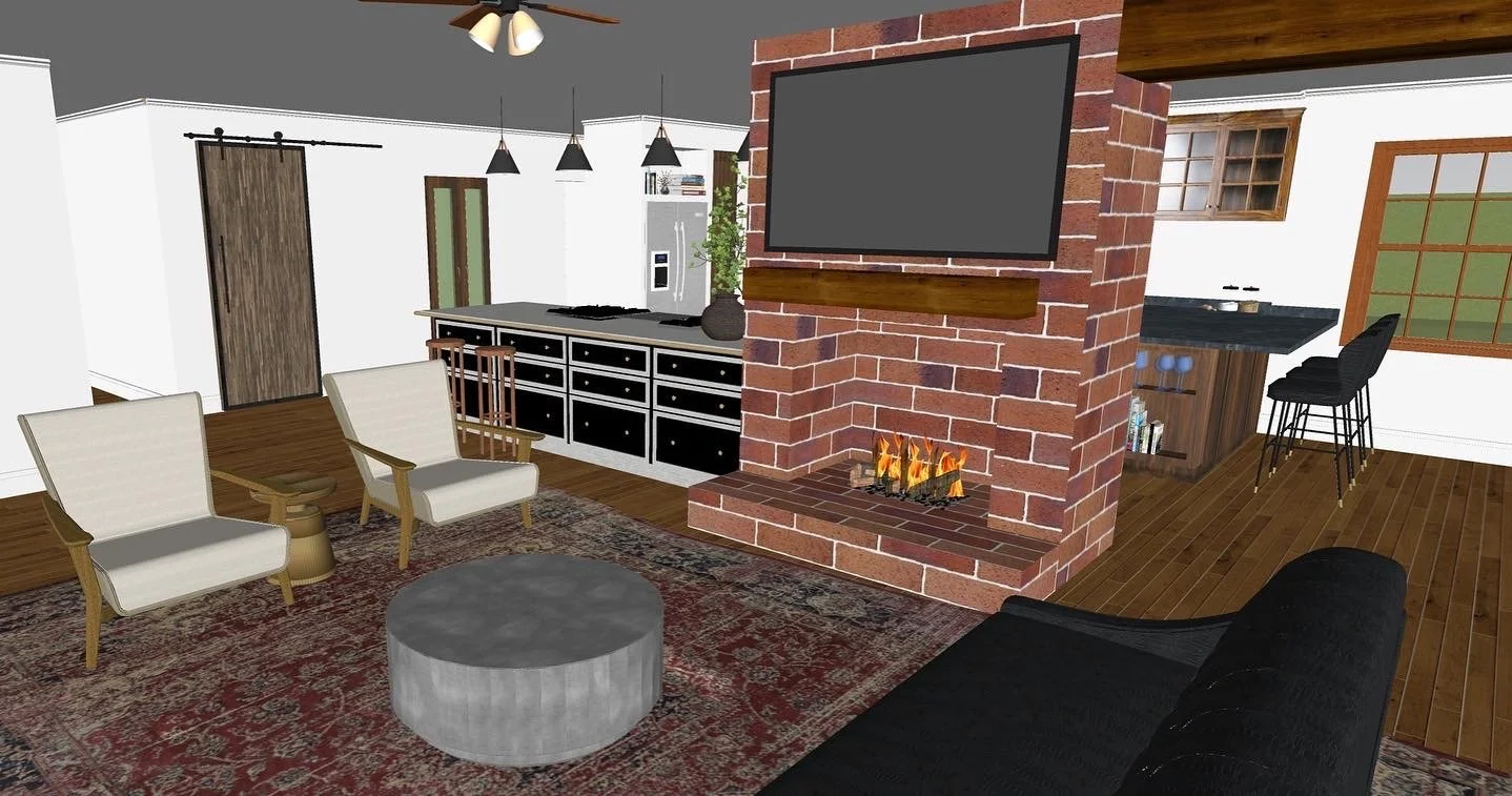 Living room with a brick fireplace, wall-mounted TV above it, two white armchairs with wooden armrests, round gray ottoman, area rug, black sofa, view into kitchen with bar stools, wooden cabinets, and a window.