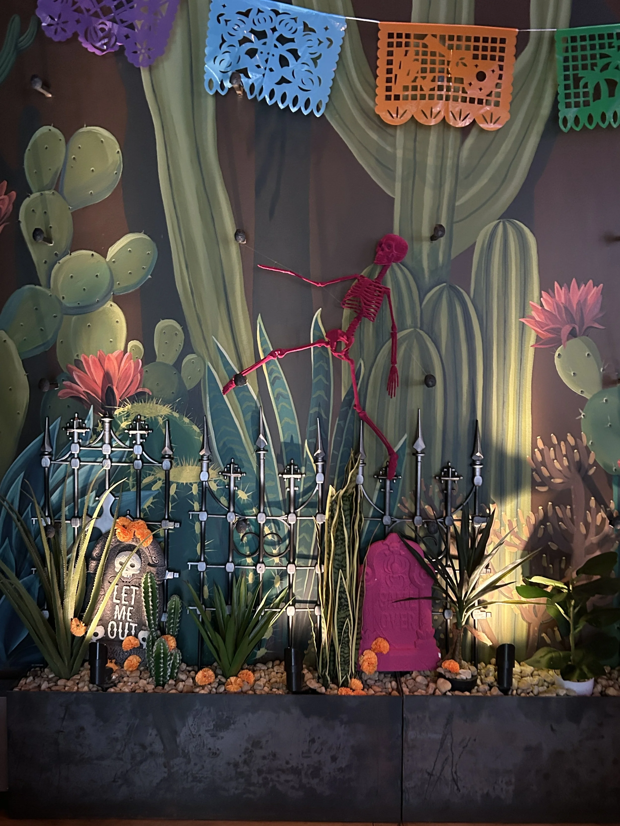 A colorful display with a painted cactus and desert scene background, decorated with Frida Kahlo-themed items, including a skeleton figure, cacti, flowers, and Mexican papel picado banners.