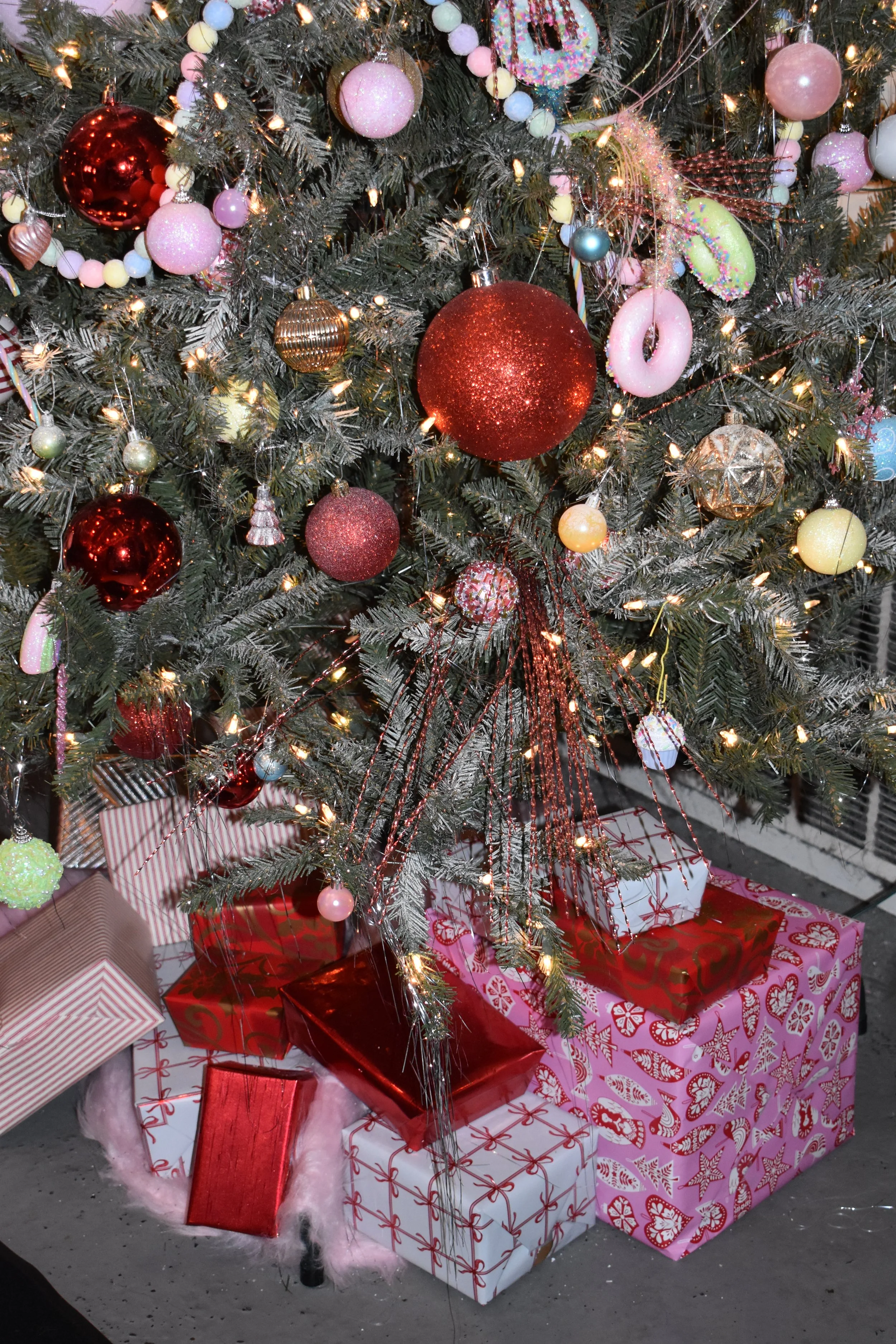 A decorated Christmas tree with pink, red, gold, and pastel ornaments and lights, surrounded by wrapped presents at the base.