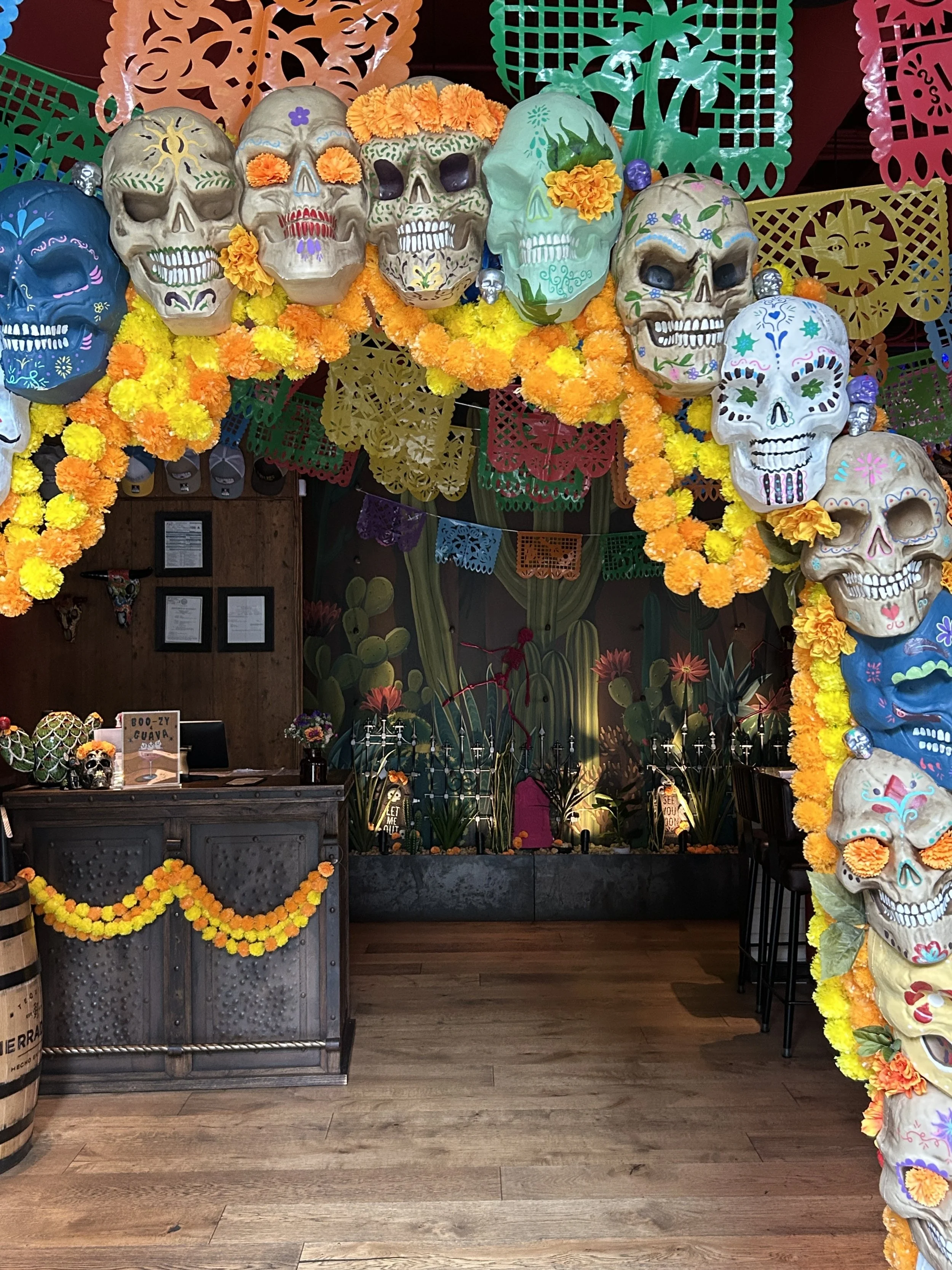 Decorative display for Day of the Dead featuring colorful skull masks, marigold flower garlands, and papel picado banners in various bright colors. Emilio's Tacos & Tequila Midtown Atlanta.