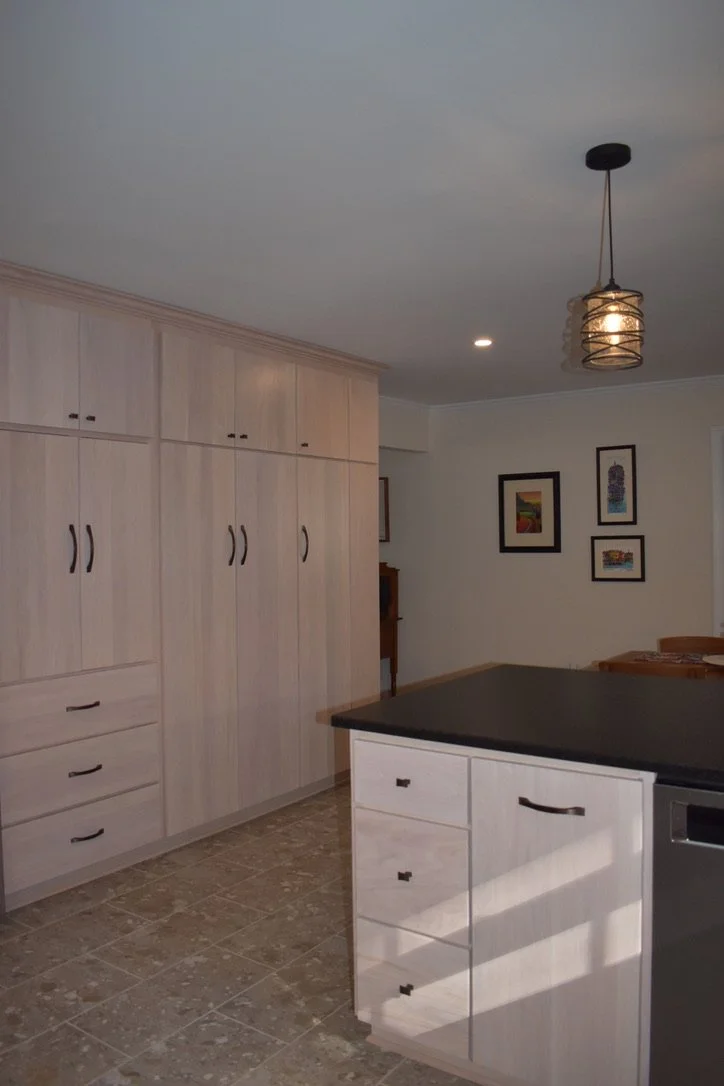 Interior of a kitchen with light-colored cabinets, a dark countertop, a small island with drawers, framed artwork on the wall, and a hanging light fixture.