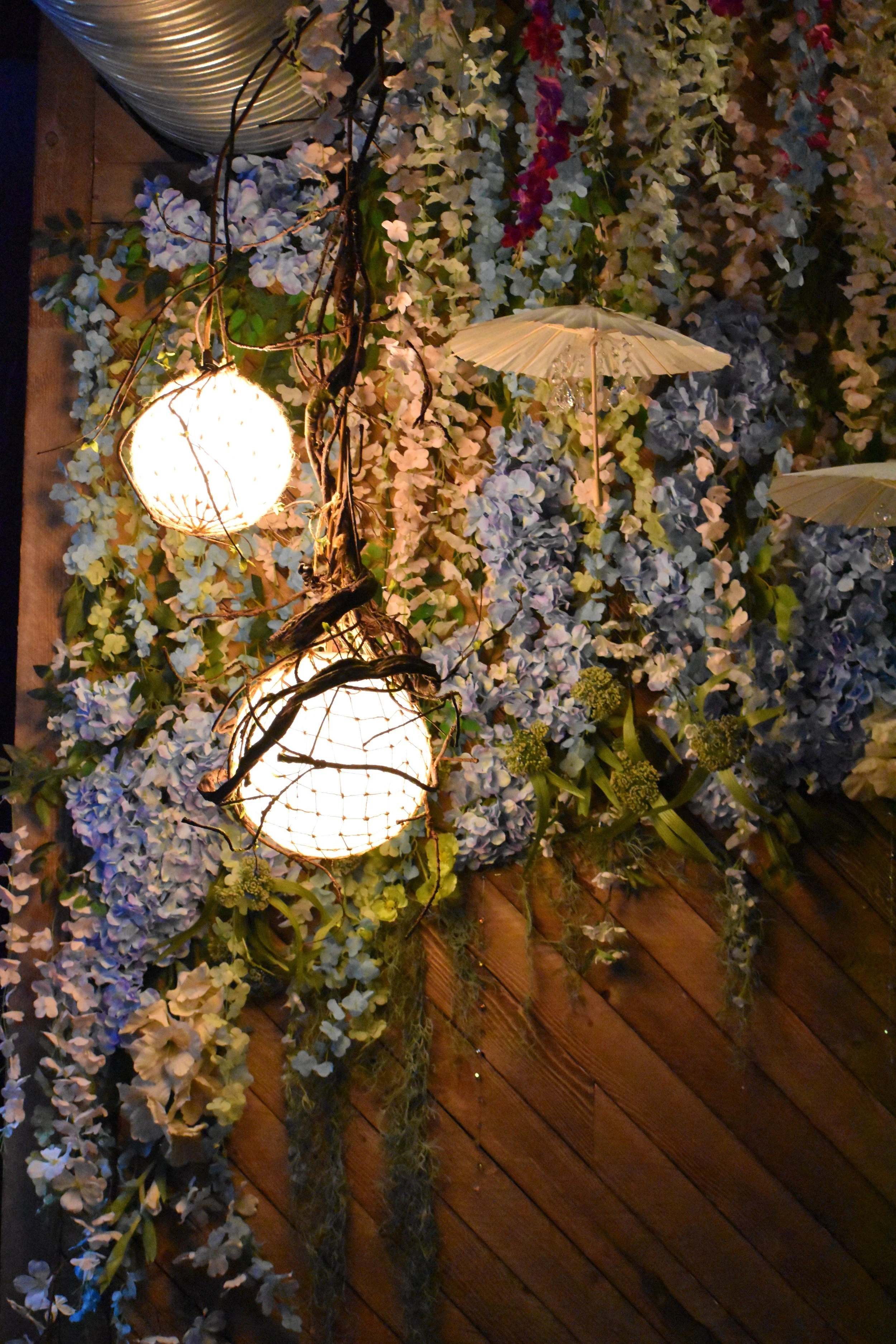 Decorative wall with hanging lights, white parasols, and cascading blue and white flowers on a wooden backdrop. Hamp & Harry's Marietta Square.