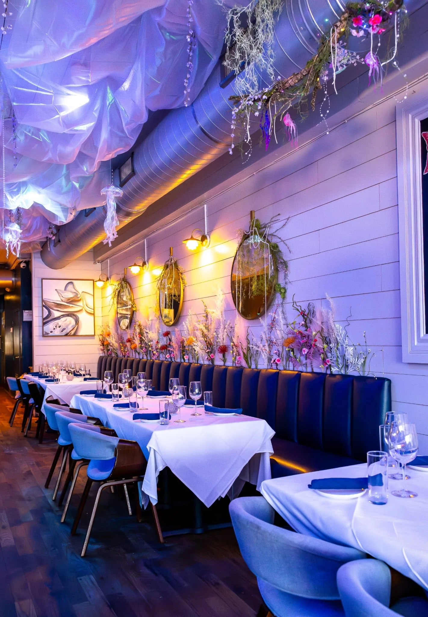 Interior of a restaurant decorated with hanging art and floral arrangements, set for dining with tables covered in white cloths, wine glasses, and blue napkins.