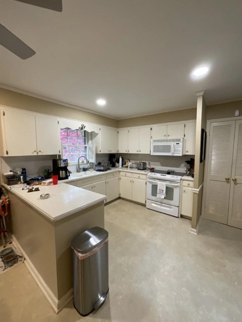A before picture of a kitchen with white cabinets, a window above the sink, a white stove, a microwave, a coffee maker, and various kitchen items on the counters. There is a trash can in the corner, and a closet with louvered doors on the right.