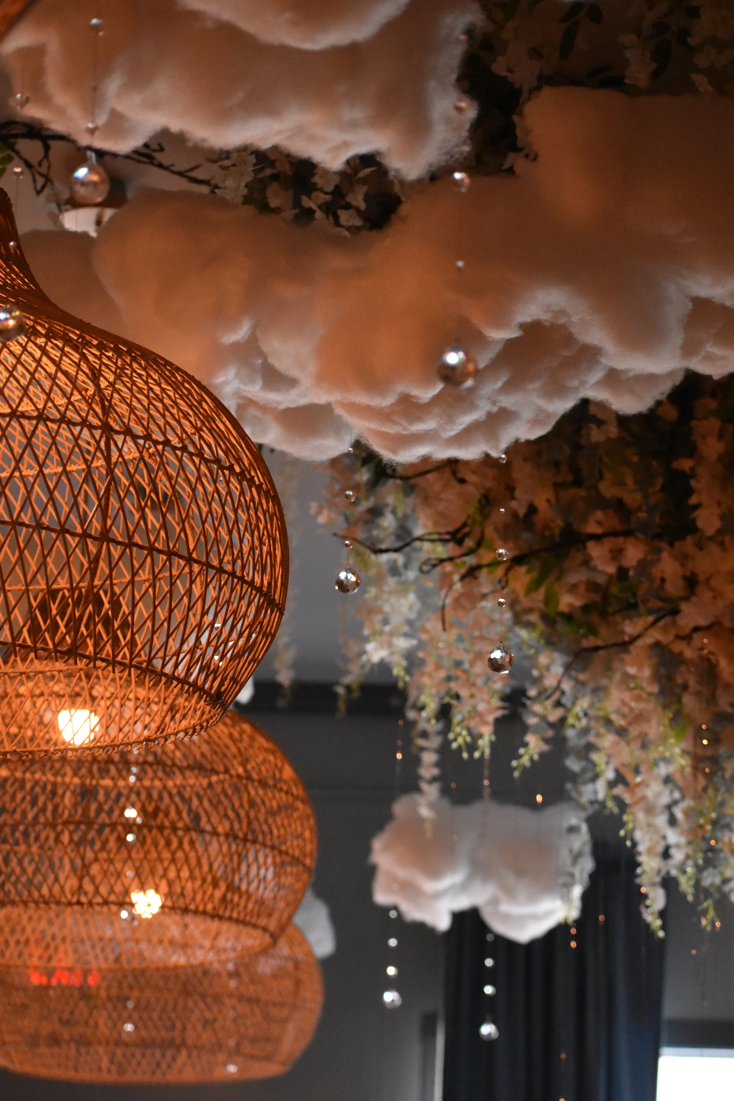 Decorative ceiling arrangement with white fluffy clouds, hanging silver beads, dried pink flowers, and illuminated orange woven pendant lights. Hamp & Harry's Marietta Square.