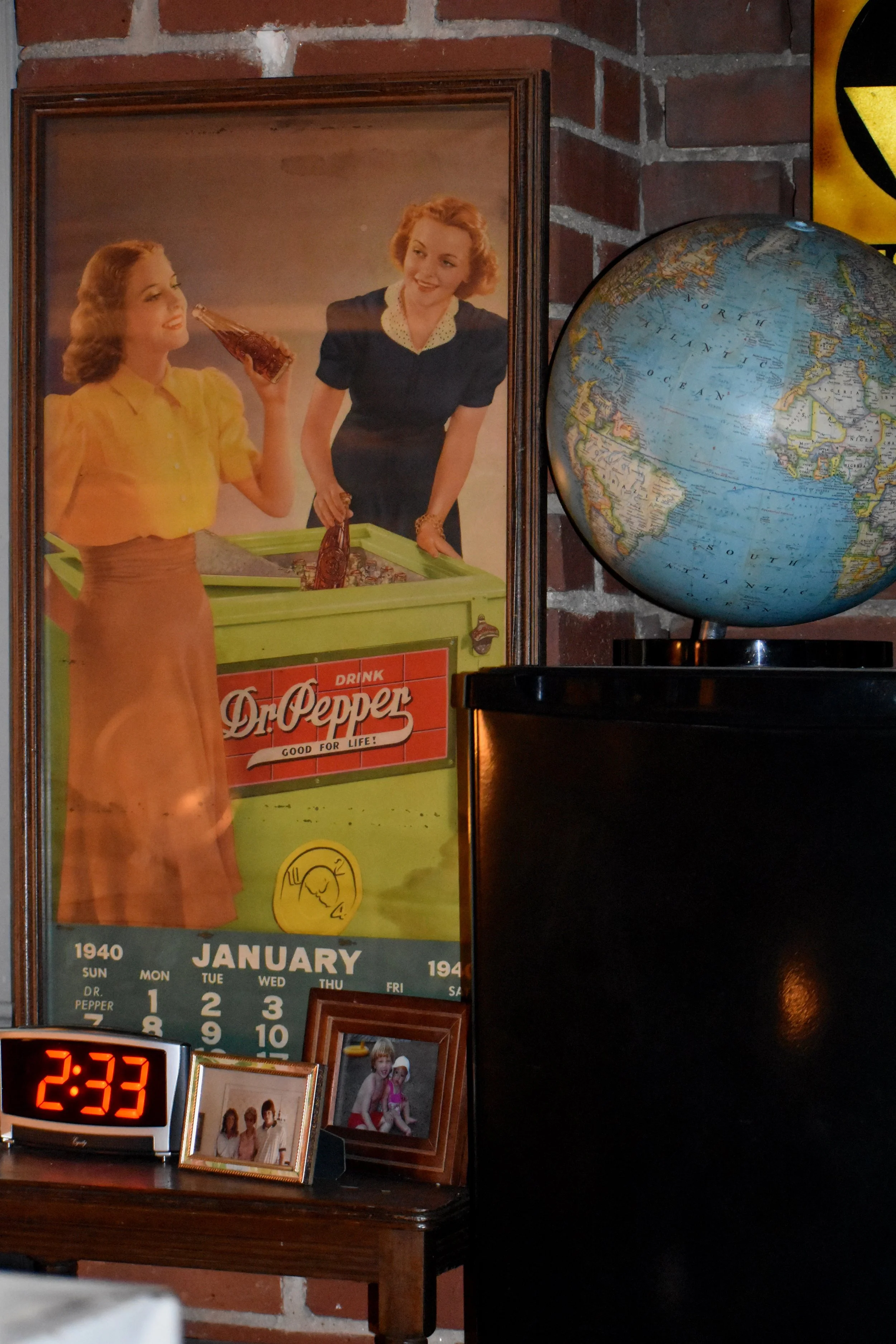 A vintage poster of two women, one holding a bottle of soda, beside a Dr. Pepper vending machine, on a brick wall. A small digital clock showing 2:33, and two framed photographs are on a wooden table, with a globe on a black stand next to the poster.