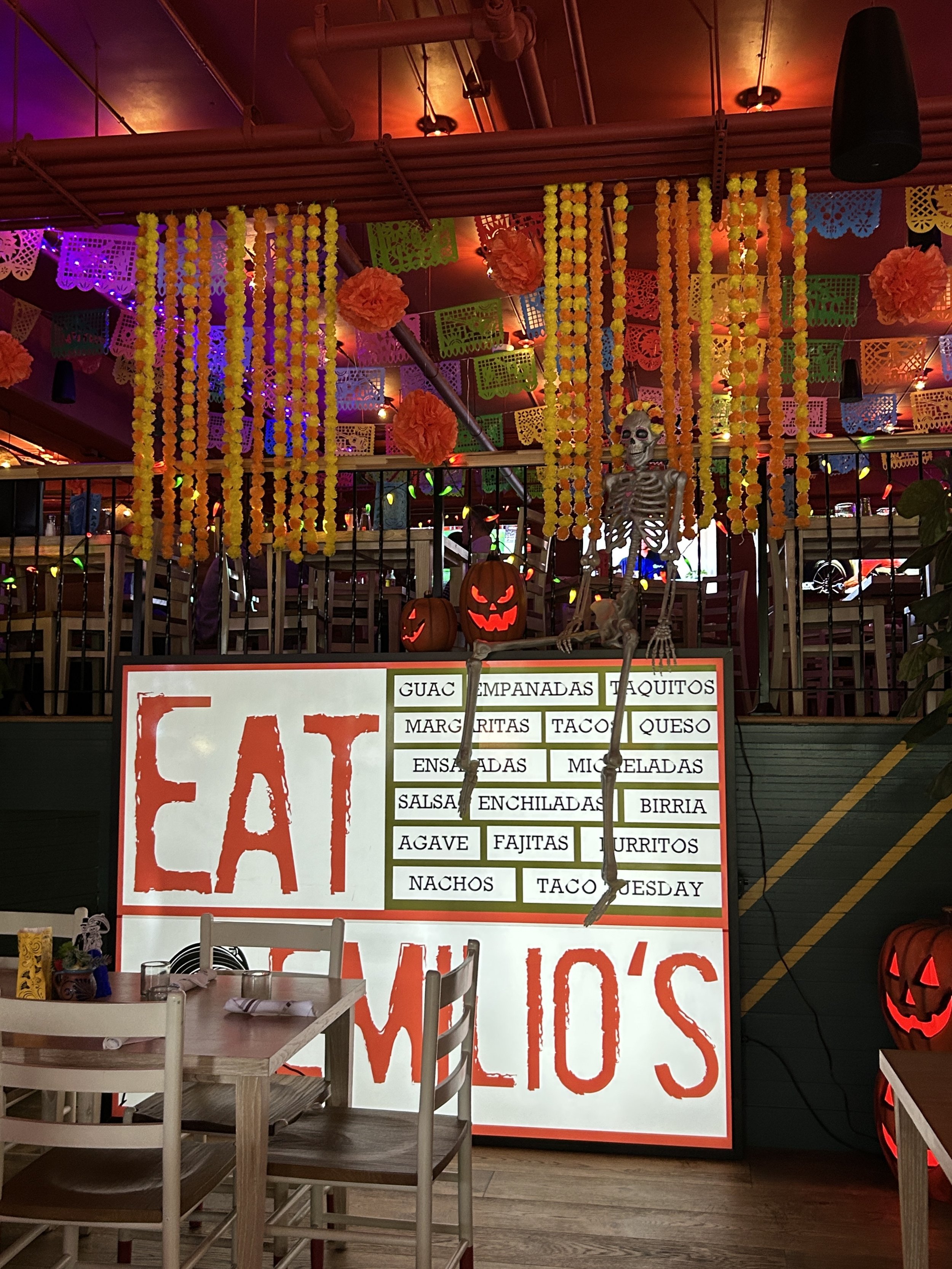 Decorated Mexican restaurant with bright hanging paper flowers and colorful papel picado banners, a skeleton hanging from the ceiling, pumpkin decorations, and a large illuminated menu sign listing Mexican dishes, with Halloween-themed decorations li