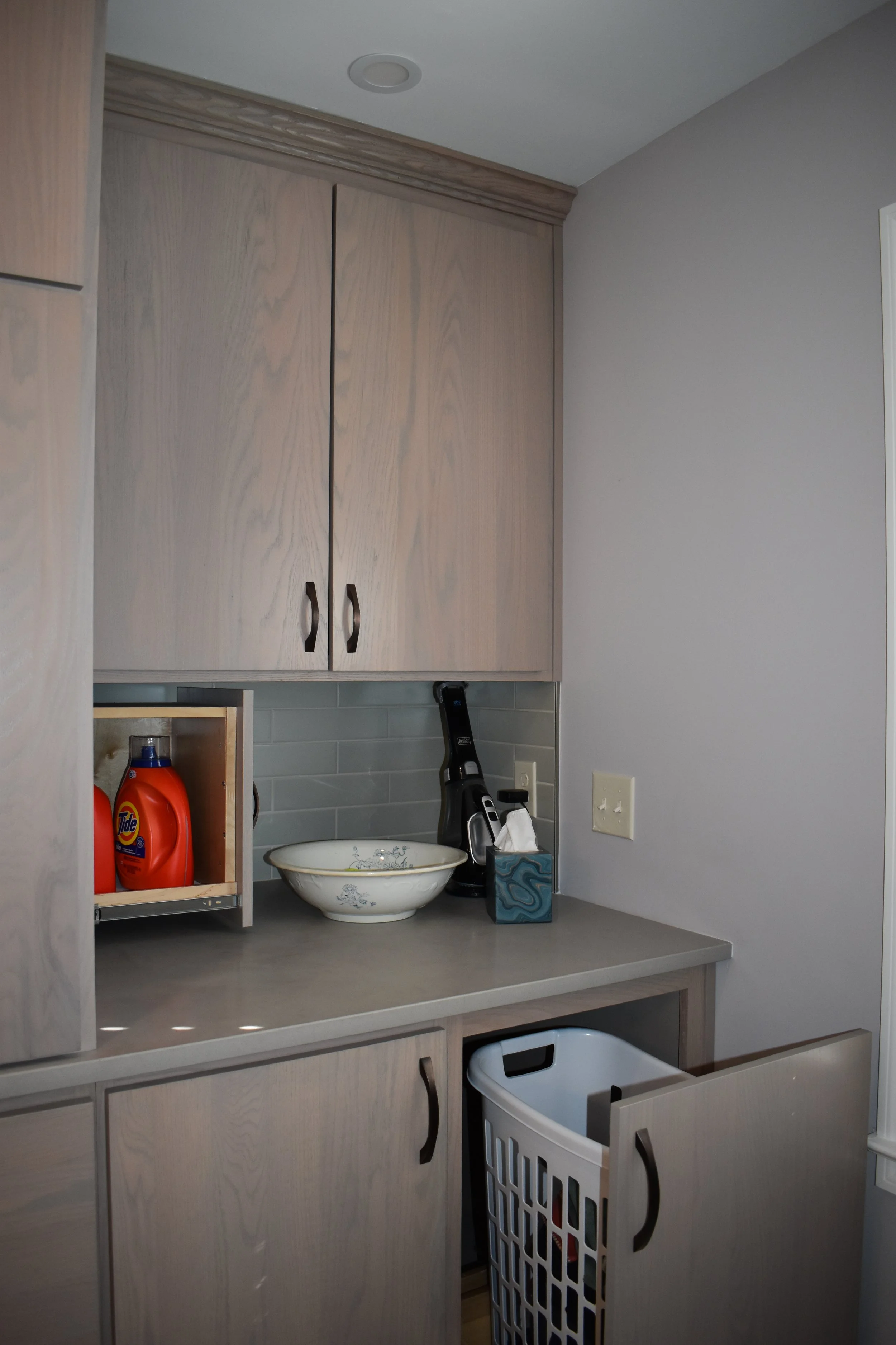 Kitchen corner with light-colored wooden cabinets, a countertop with a bowl, dryer sheets, laundry detergent, and a handheld vacuum, and a plastic laundry basket. Marietta GA bathroom remodel.