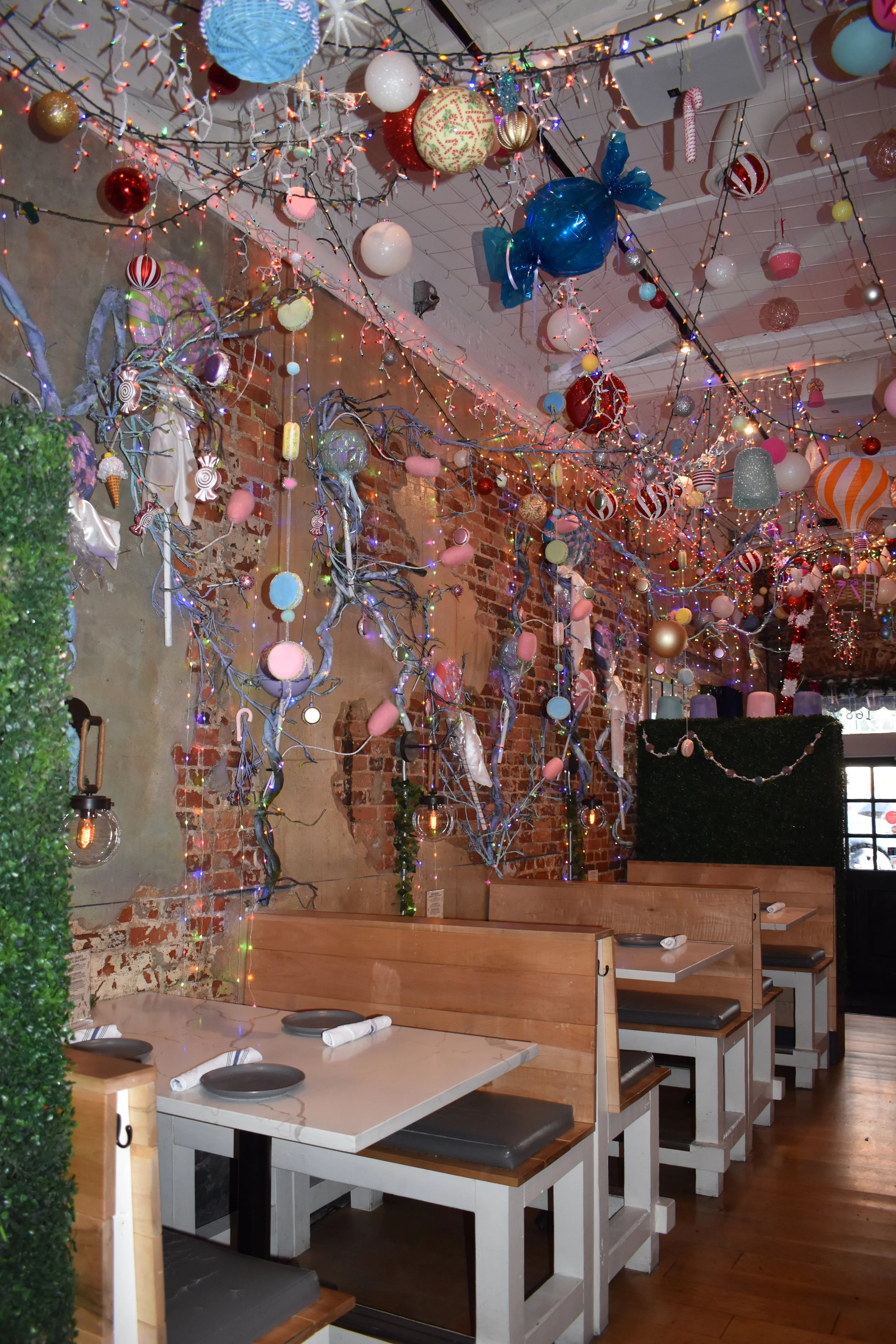 Interior of a restaurant decorated for Christmas with hanging ornaments, string lights, and festive decorations on exposed brick walls and ceiling, with wooden booths and tables set for dining.