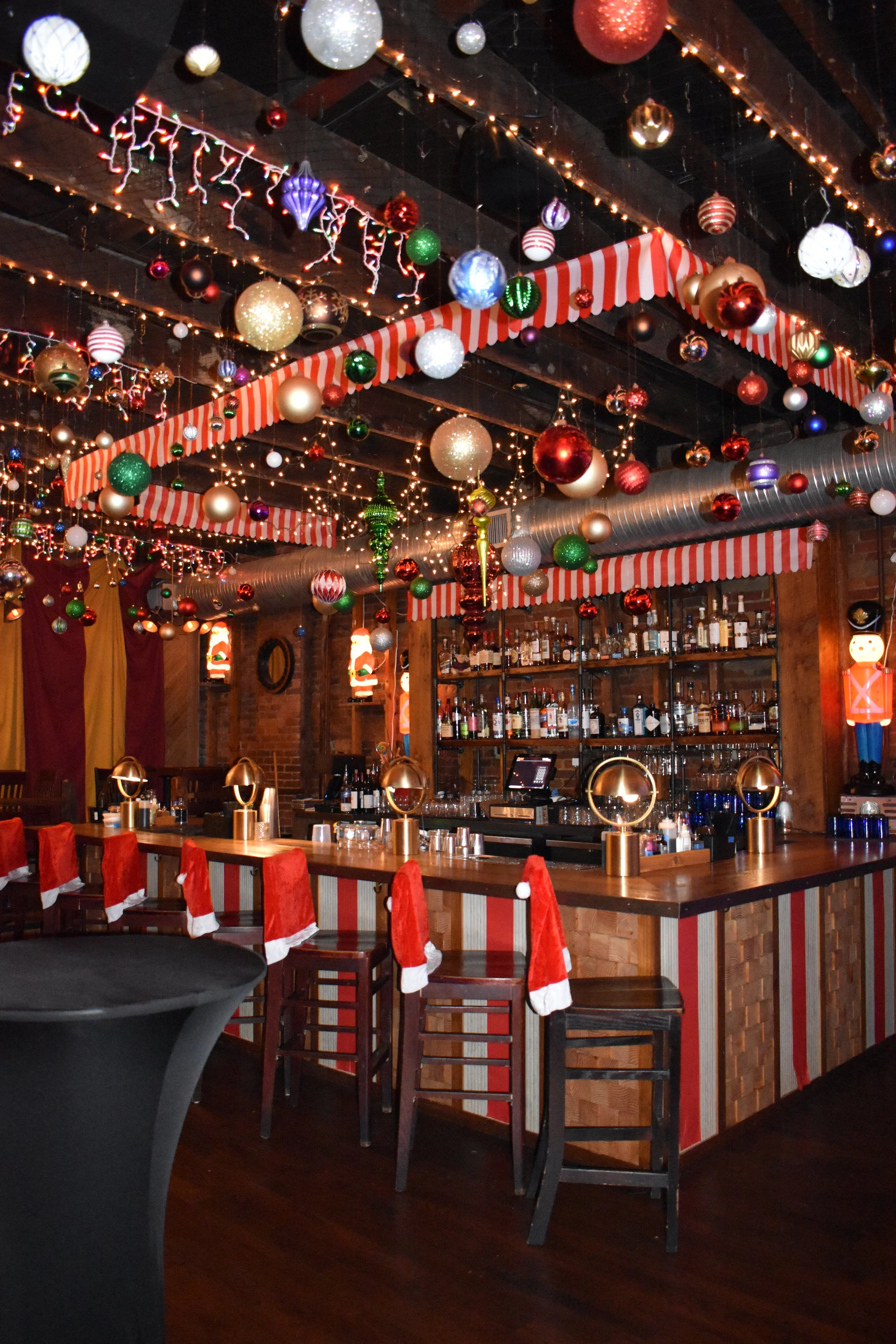 Bar decorated for Christmas with hanging ornaments, string lights, and festive red and white striped curtains. Bar stools are adorned with Santa hats, and shelves behind the bar are stocked with bottles.