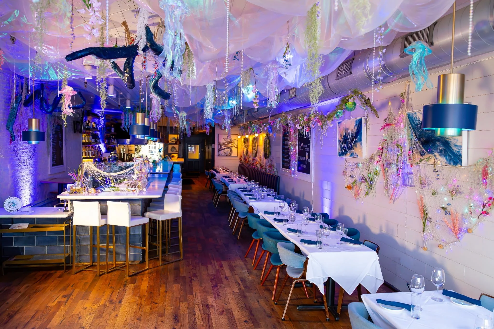 Decorated indoor restaurant with hanging fabric, string lights, and sea-themed ornaments, featuring long tables with white and blue chairs, and a bar area in the background.