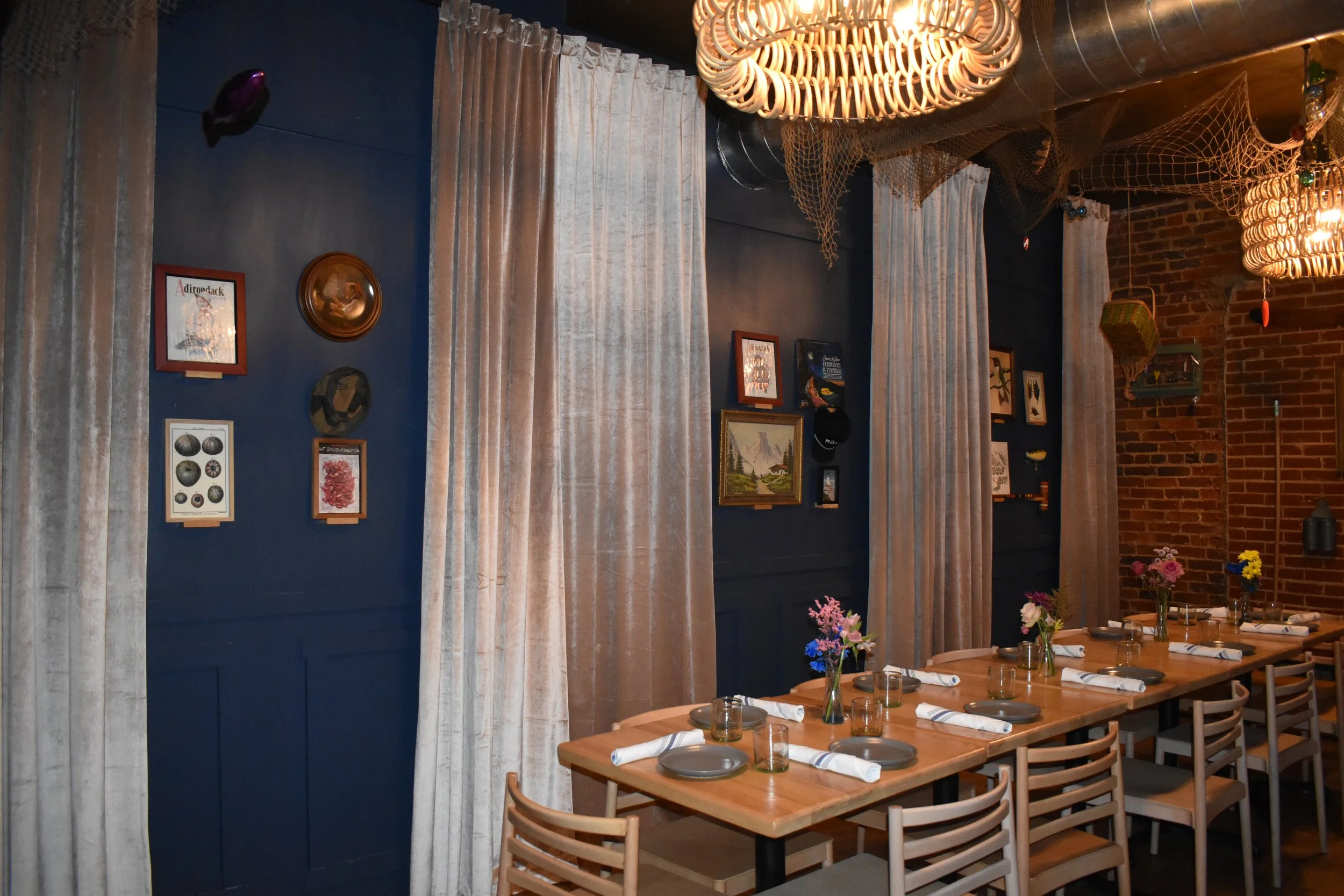 Interior of a restaurant with a long wooden table set with plates, glasses, napkins, and small flower arrangements. The background features a brick wall and dark blue painted wall decorated with framed pictures, plates, and hats. Cream-colored curtai