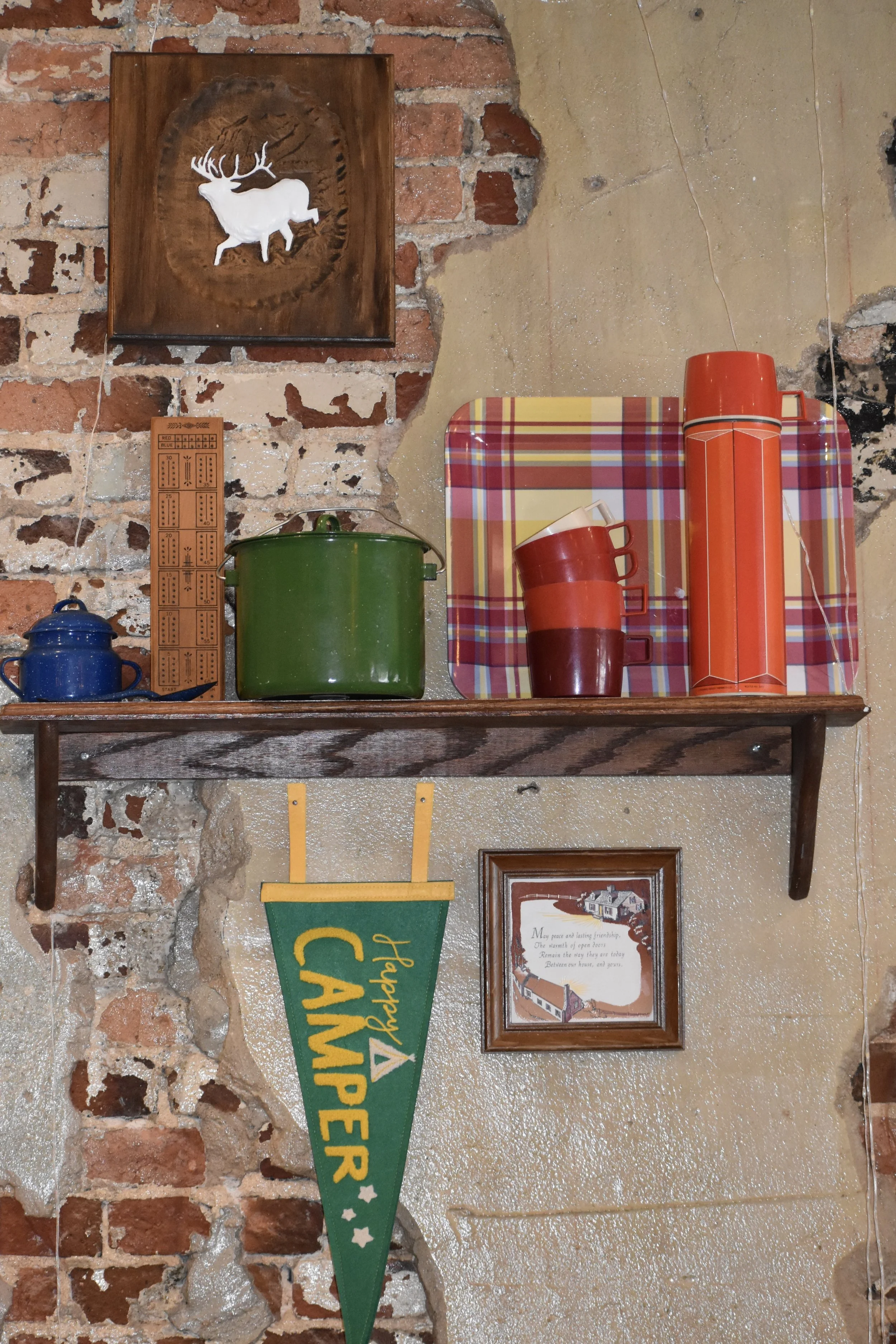 A rustic kitchen shelf with colorful cookware and vintage decor, including a framed picture of a house and a green camping pennant. Hamp & Harry's Marietta Square.