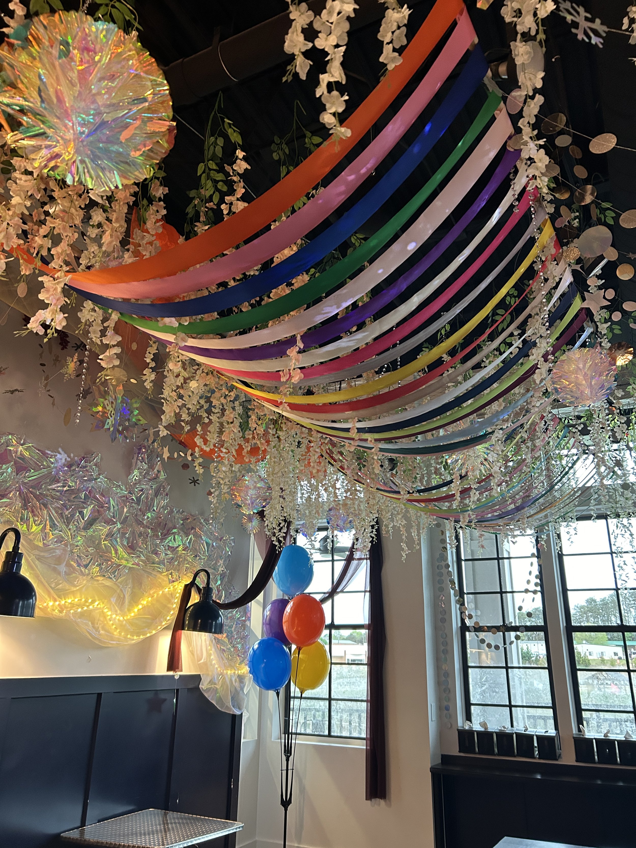Colorful party decorations hanging from the ceiling with rainbow streamers, white floral garlands, balloons, fairy lights, and reflective hanging accents in a room with windows.