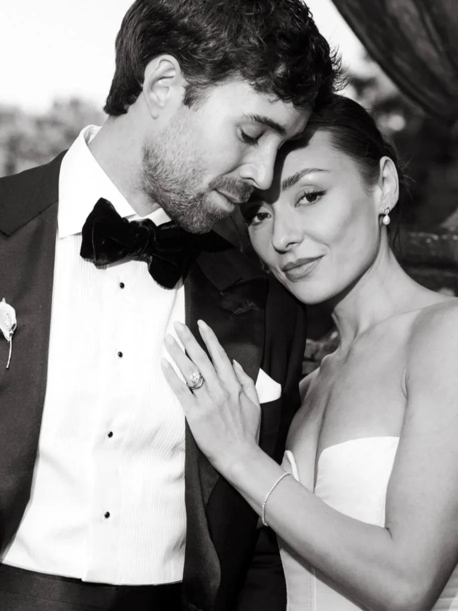 A black-and-white close-up photo of a bride and groom, with the groom in a tuxedo and the bride in a strapless dress. They are touching foreheads and smiling softly, with the bride's hand on the groom's chest showing her wedding ring.