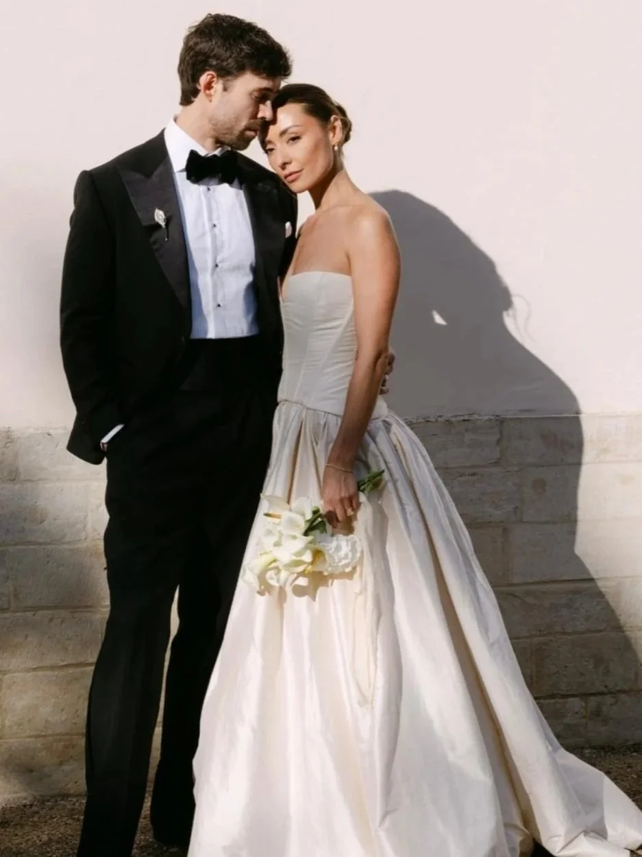 A bride and groom pose together at their wedding, with the bride holding a small bouquet of white flowers and wearing a white strapless wedding gown, and the groom in a black tuxedo with a black bowtie, against a plain wall background.