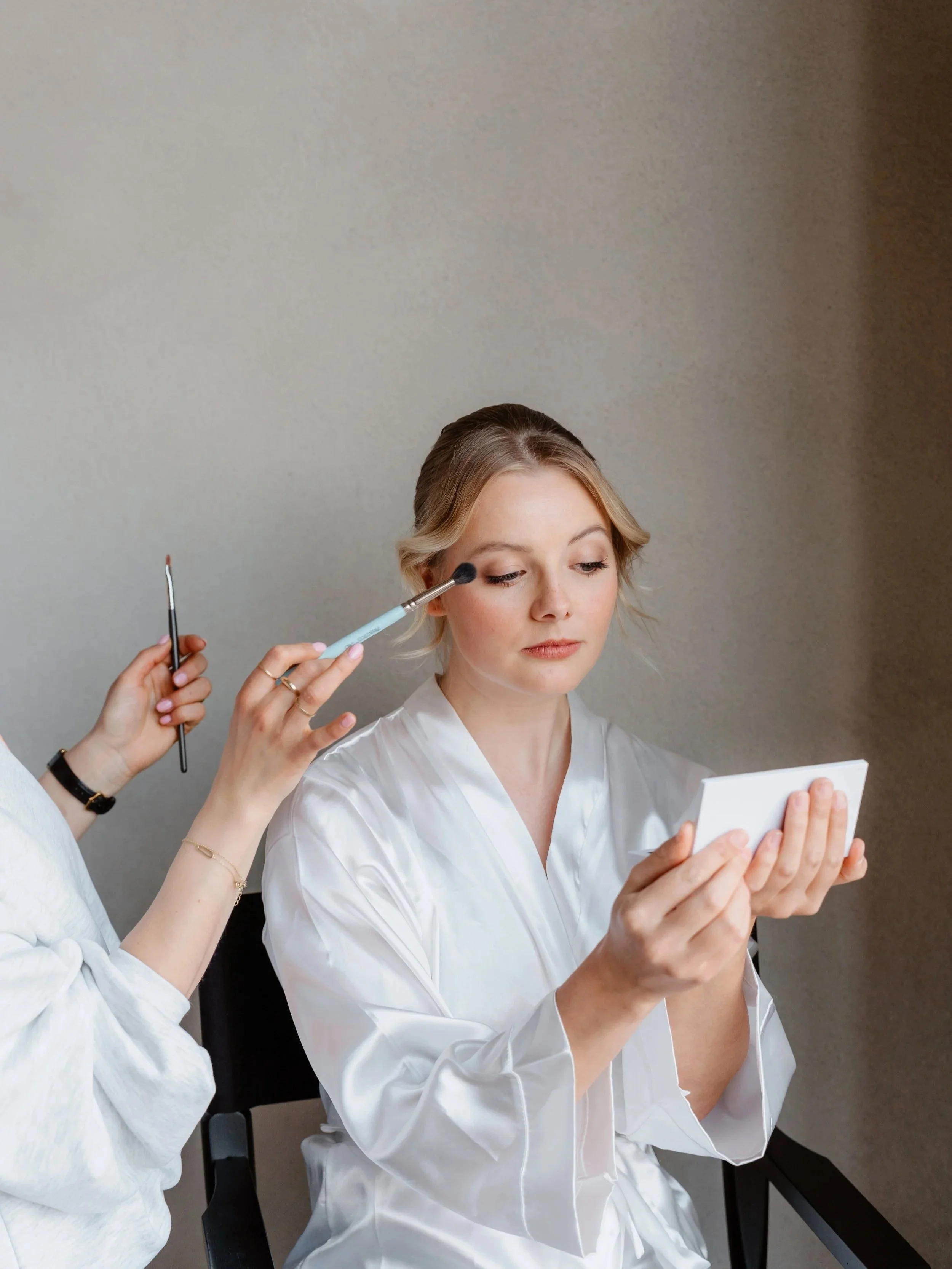 A woman in a satin robe is applying makeup, holding a mirror in her hand. A makeup artist is using a brush to apply eyeshadow or blush to her face.