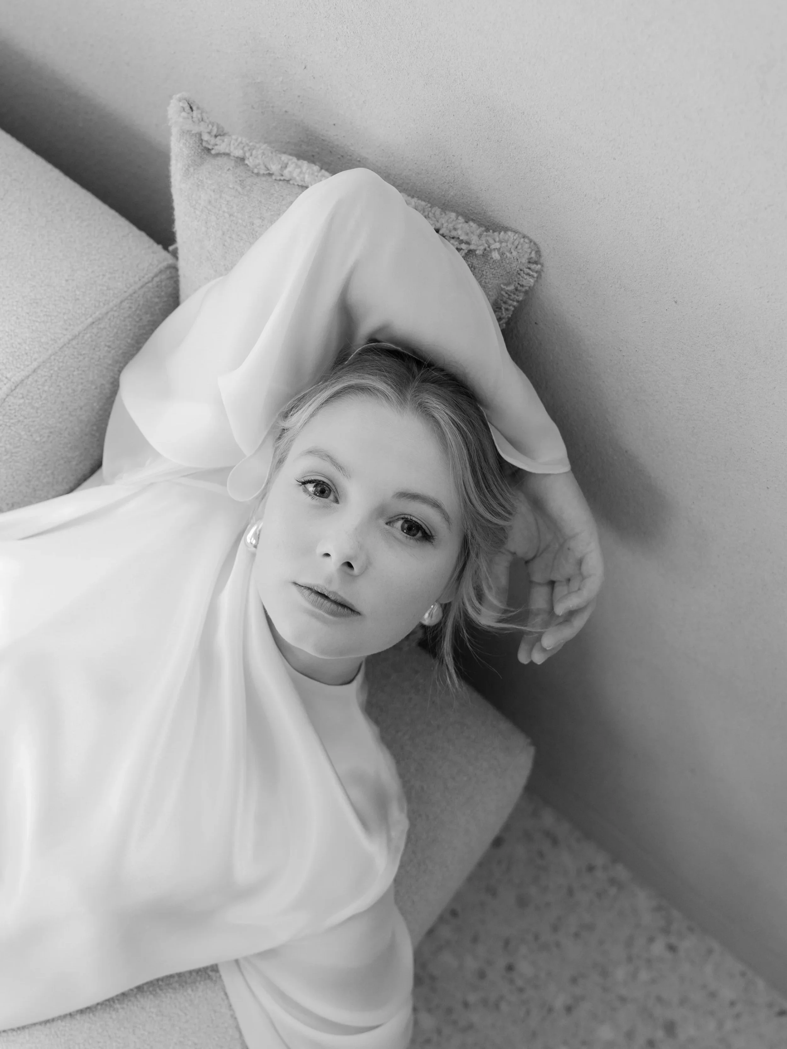 A black and white photograph of a woman lying on a couch, with her head resting on a pillow. She has light-colored hair and is wearing pearl earrings and a light-colored, high-neck blouse. She gazes directly at the camera with a calm expression.