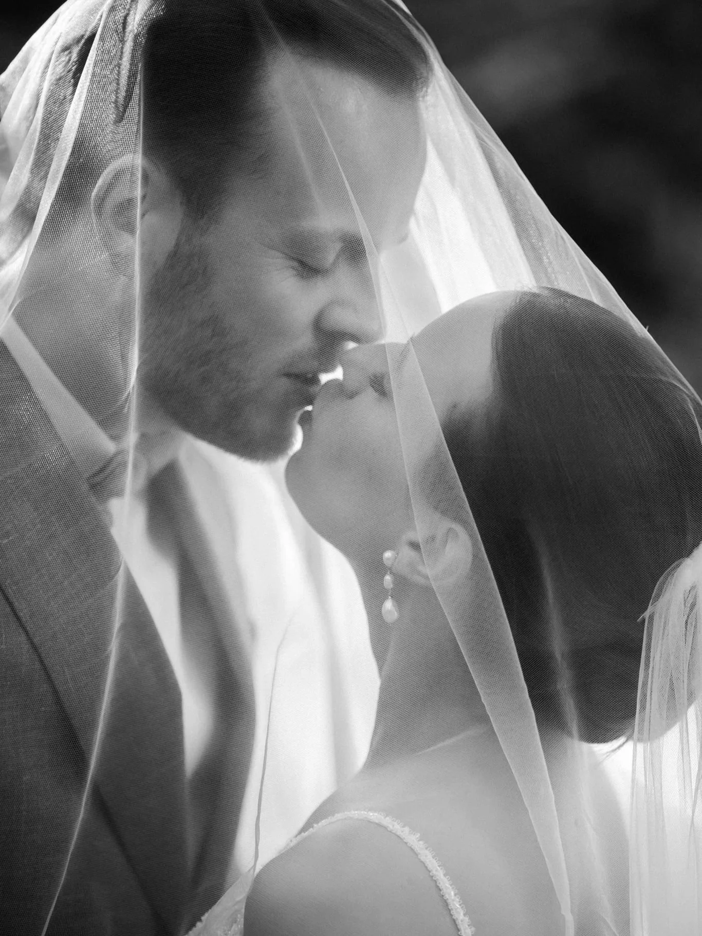 A black and white photograph of a bride and groom sharing a kiss, with the bride's face partially obscured by her veil.