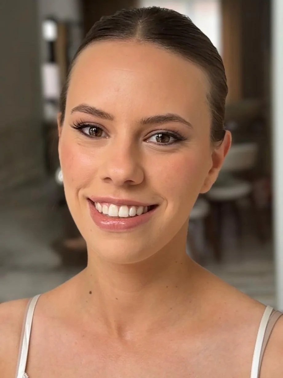 Close-up of a smiling woman with brown hair in a bun, wearing a light-colored sleeveless top, in a home setting.