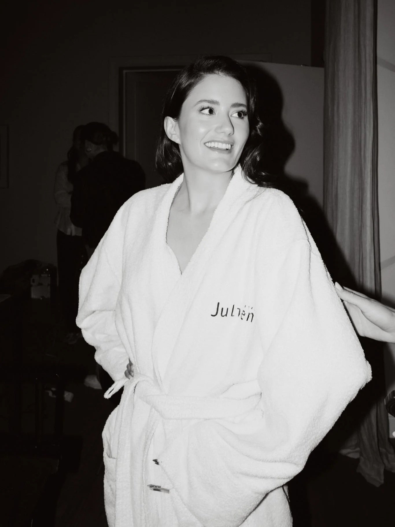 A woman with shoulder-length dark hair smiling while wearing a white bathrobe with embroidered name 'Julien' on it, standing indoors in front of a curtain and dark wall.