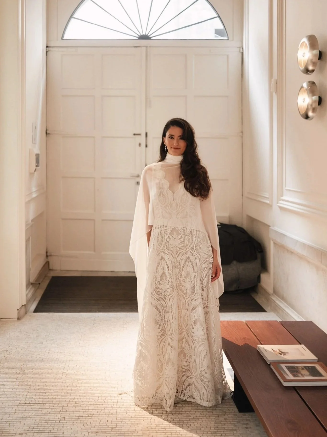 A woman in a white lace dress standing in a brightly lit entryway next to a wooden table with magazines.