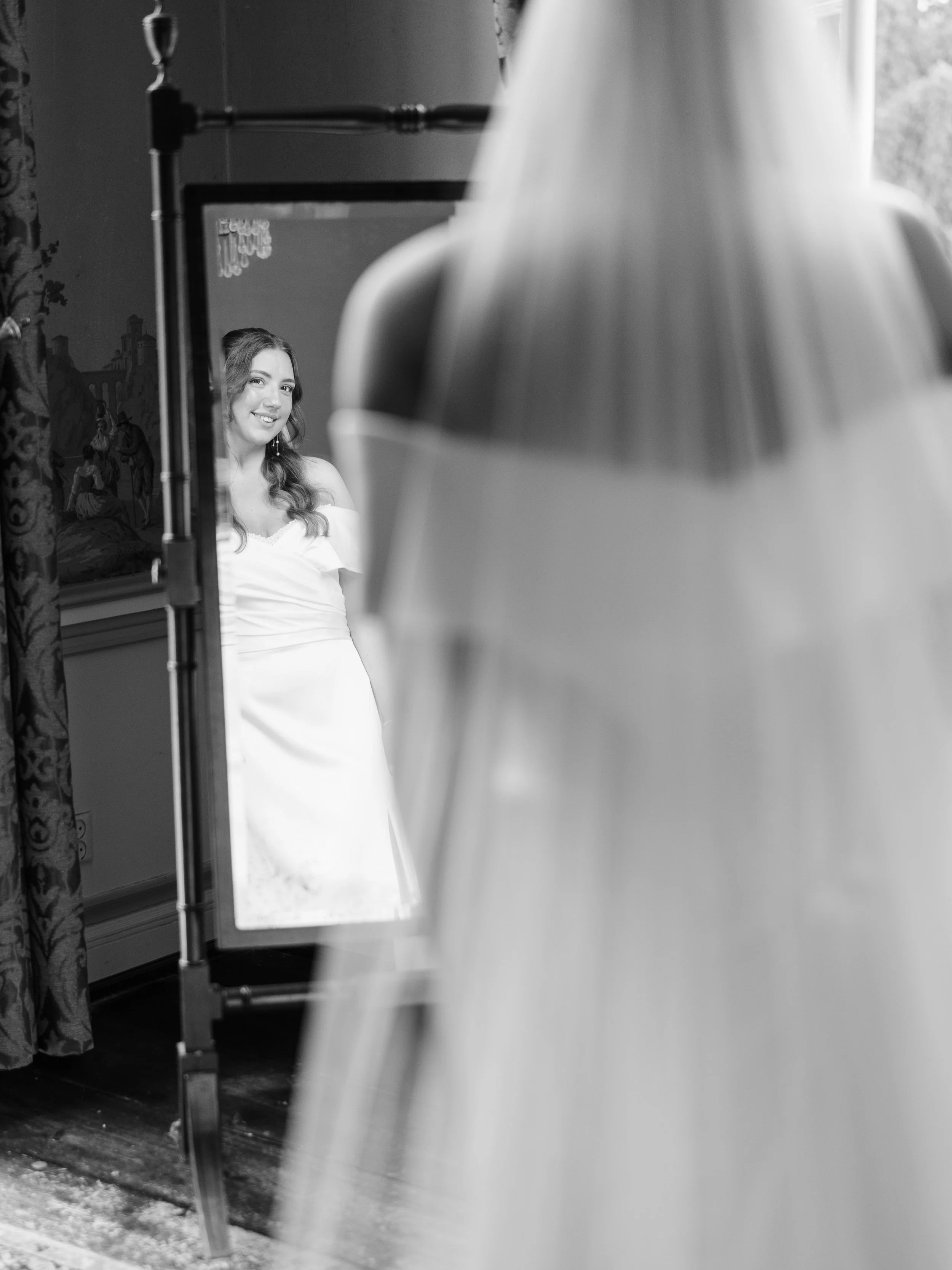 A woman in a white dress looks at herself in a mirror, smiling.
