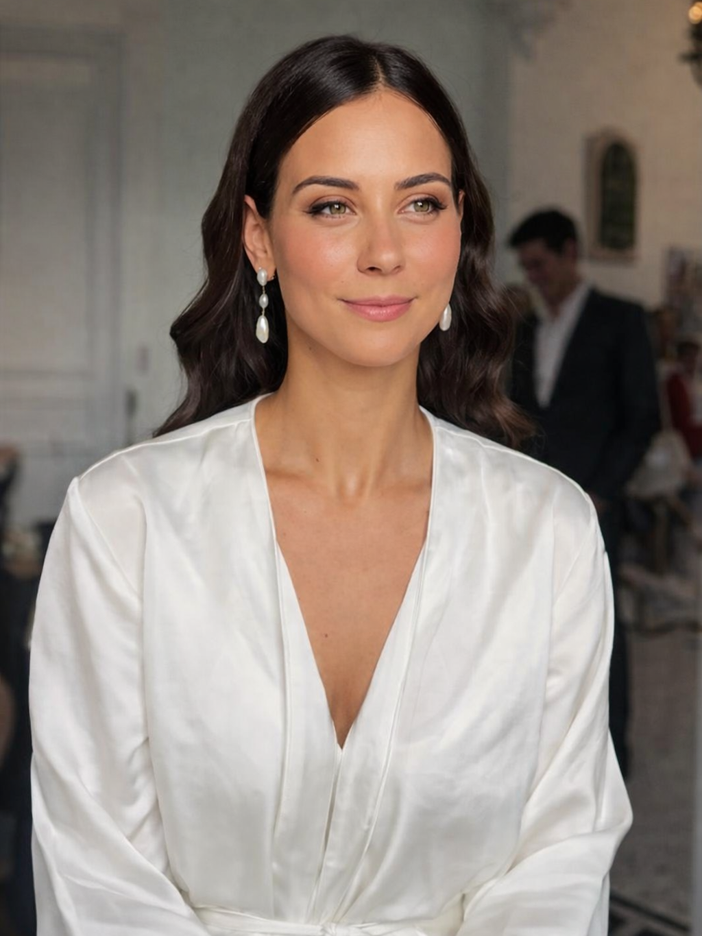 A woman with long dark hair and light makeup, wearing pearl earrings and a white satin blouse, smiling softly, indoors at a social event.