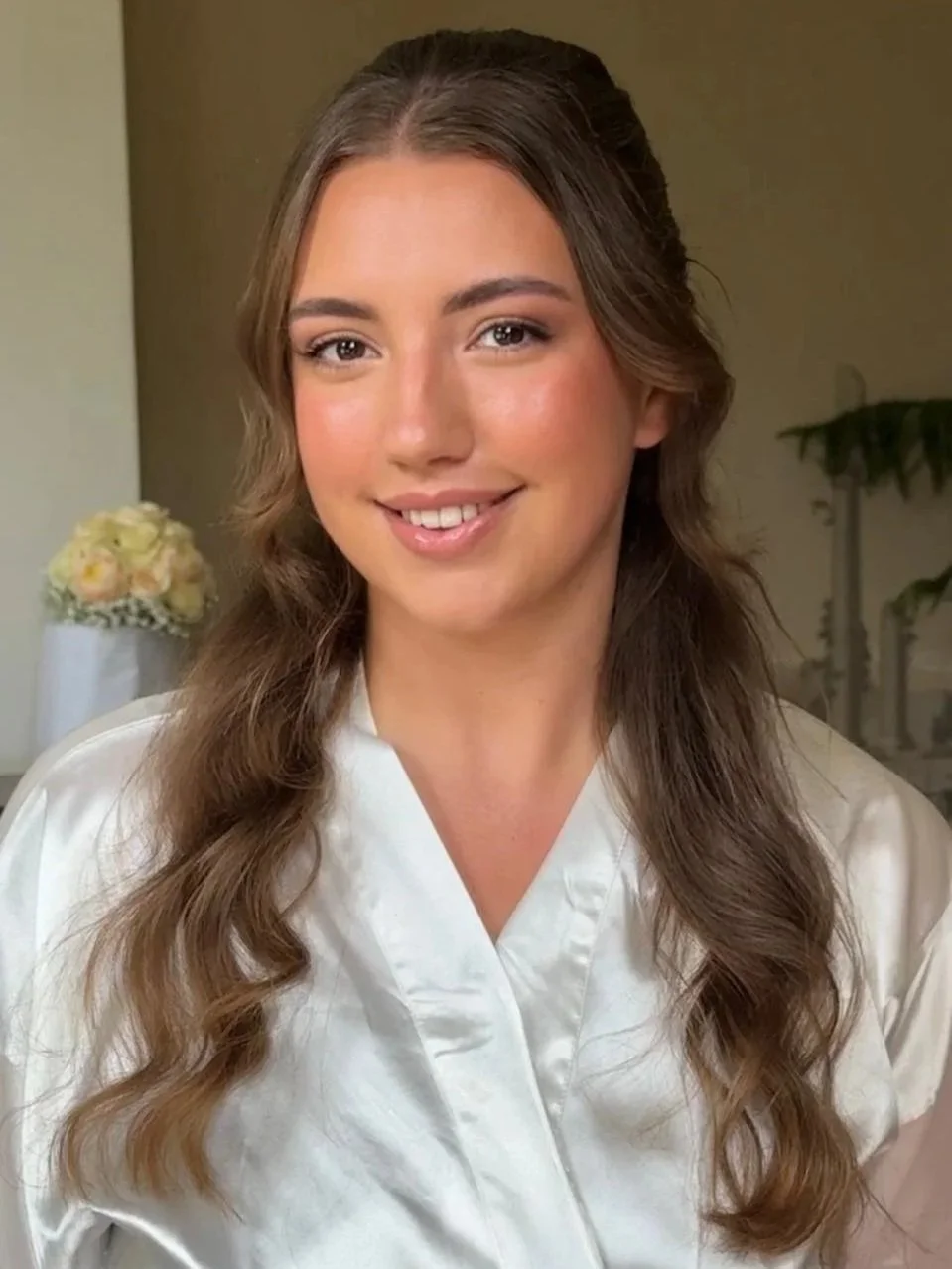 A young woman with long, wavy brown hair smiling, wearing a white satin robe, in a softly lit room with a blurred background that includes a potted plant and a flower arrangement.